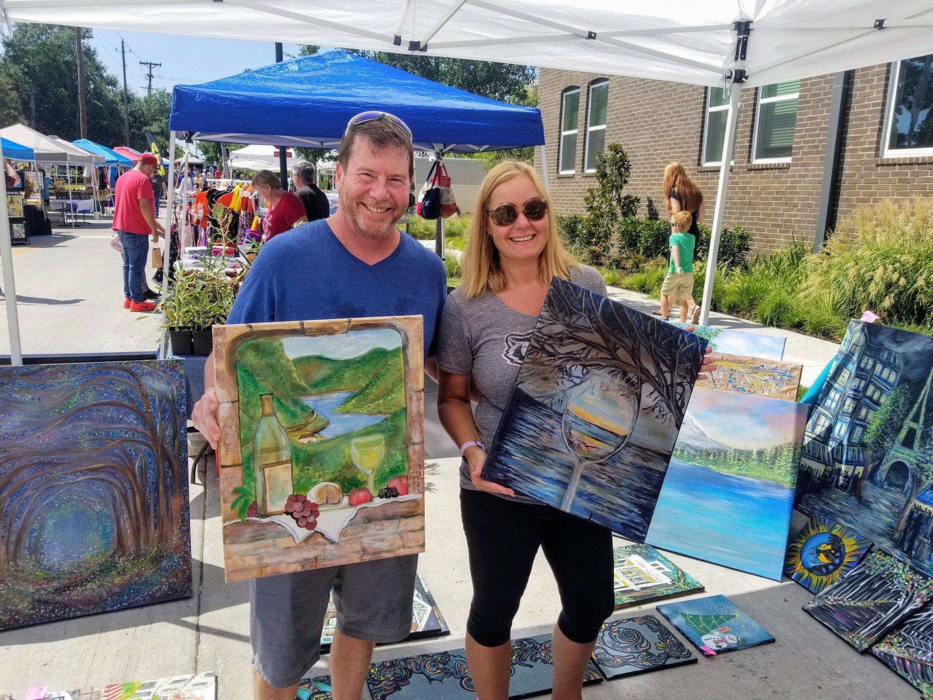 A man and a woman are holding paintings in front of a tent.