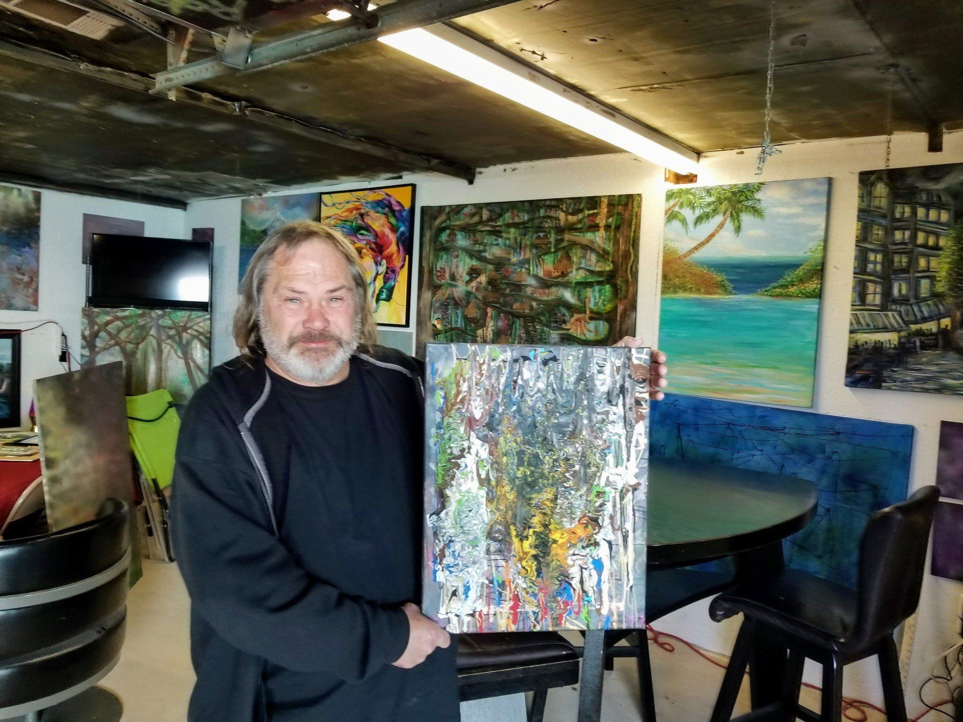 A man is holding a painting in a room with paintings on the wall.