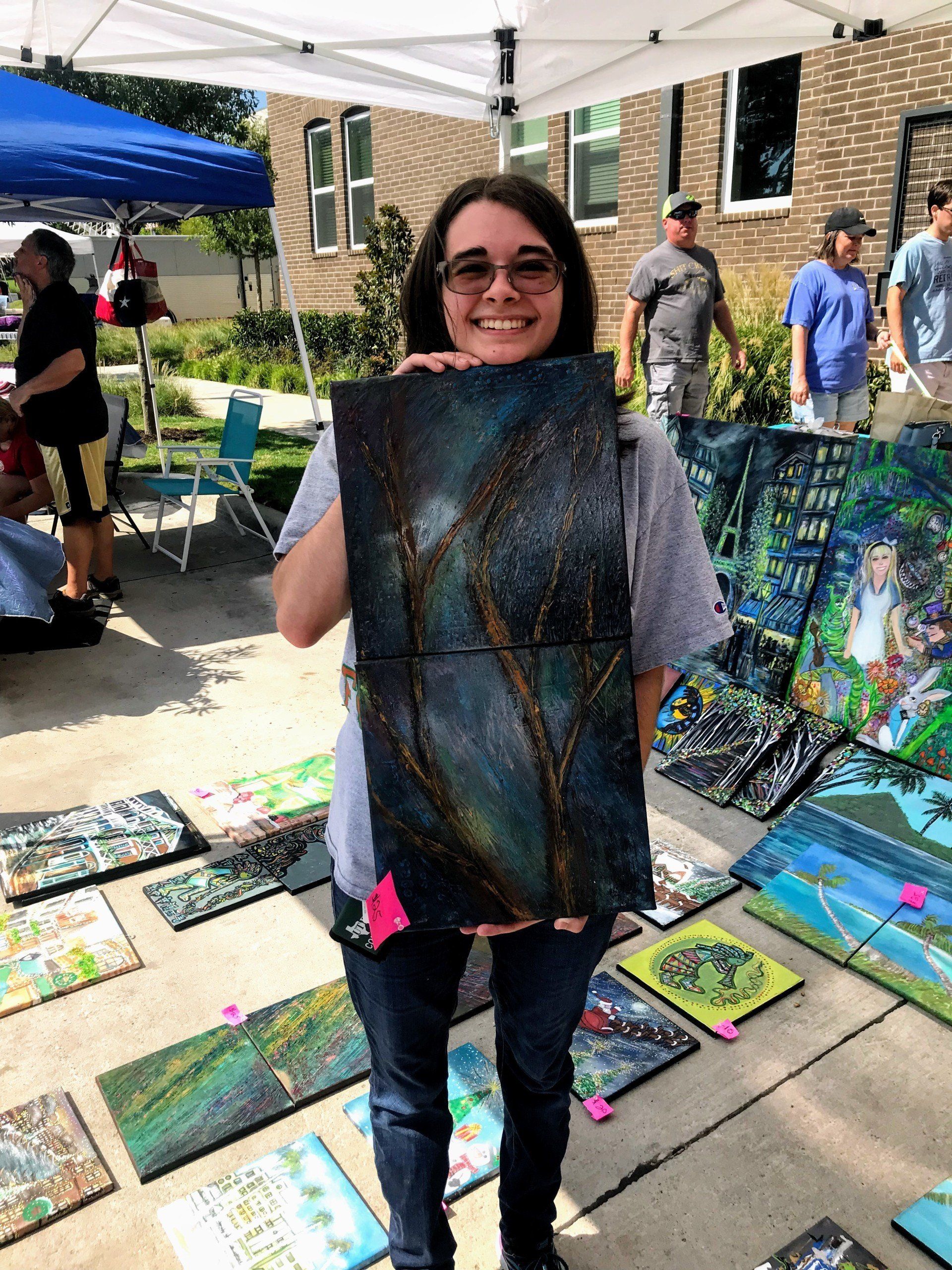 A woman is holding a painting in front of a tent.