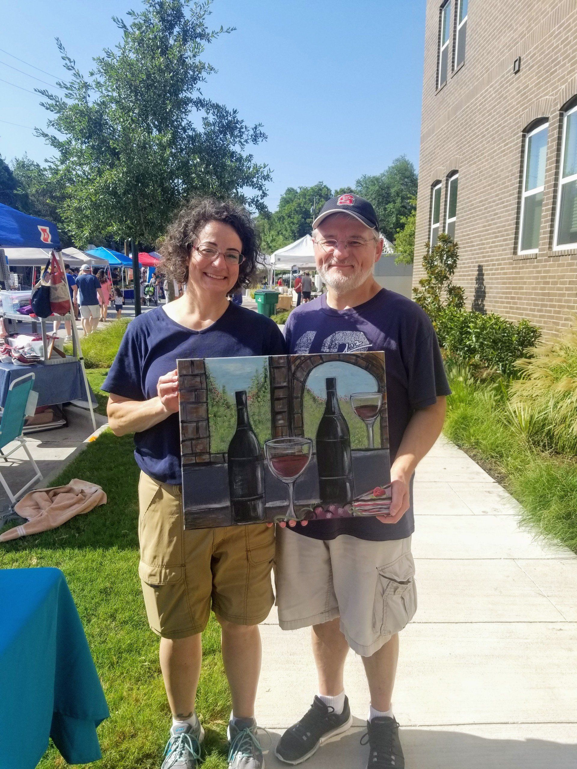 A man and a woman are standing next to each other holding a painting of wine bottles and glasses.