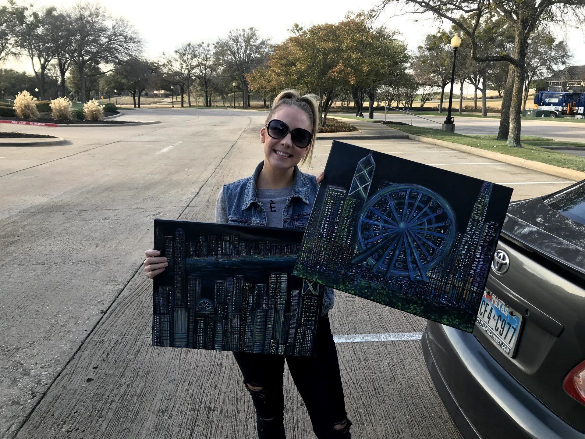 A woman is holding two paintings in front of a car.