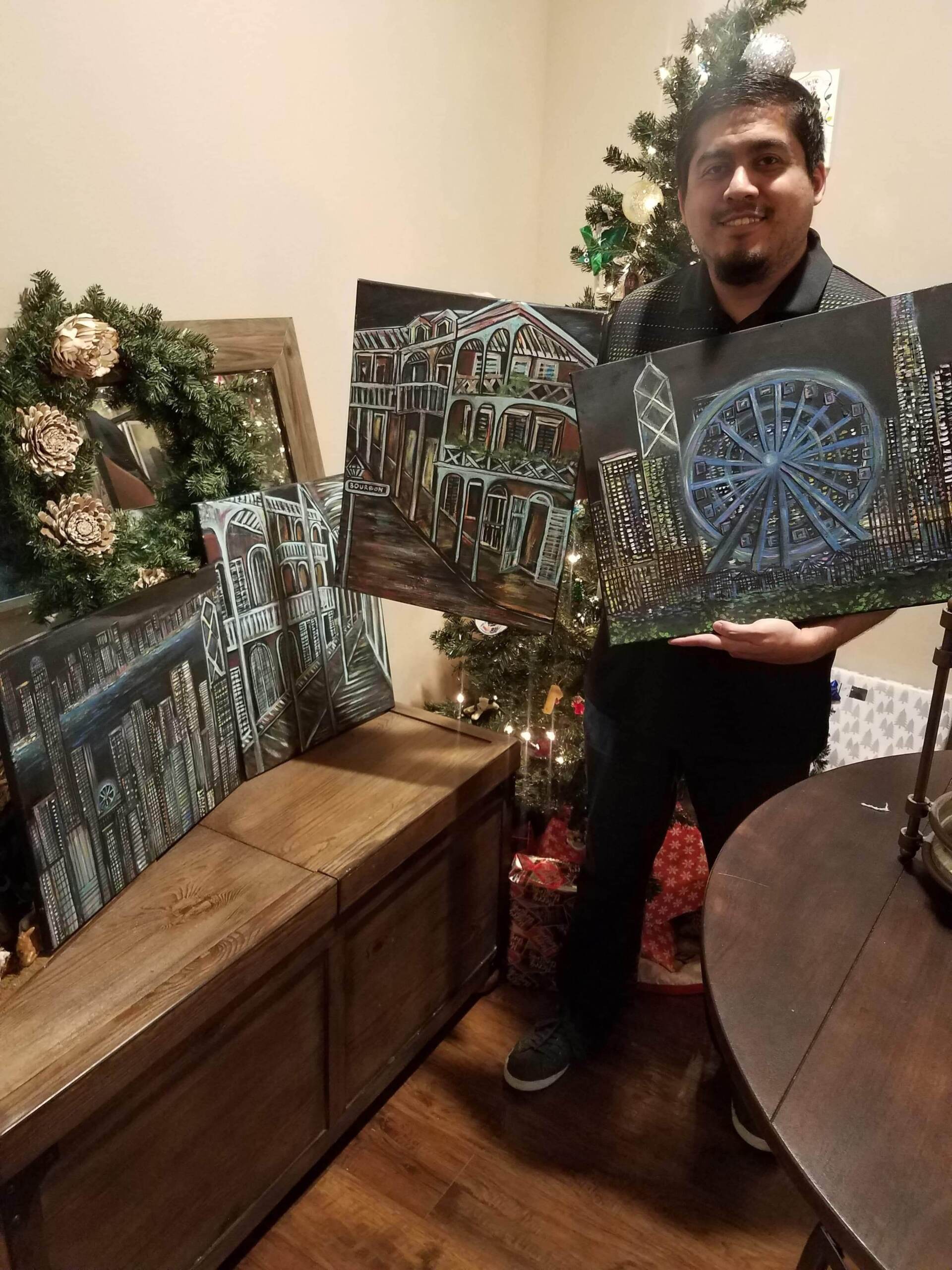 A man is holding a painting of a ferris wheel in front of a christmas tree.