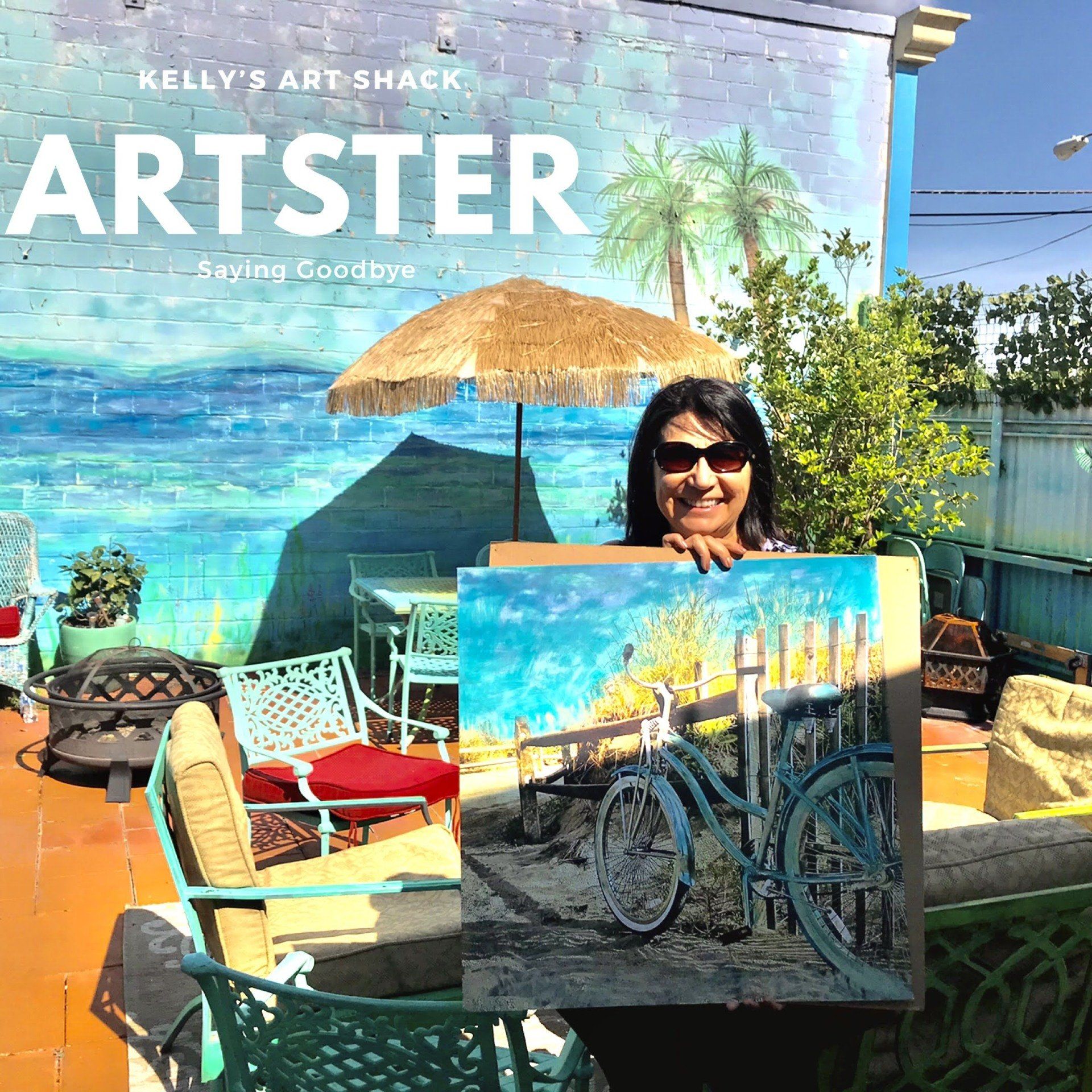 A woman is holding a painting in front of a wall that says kelly 's art shack
