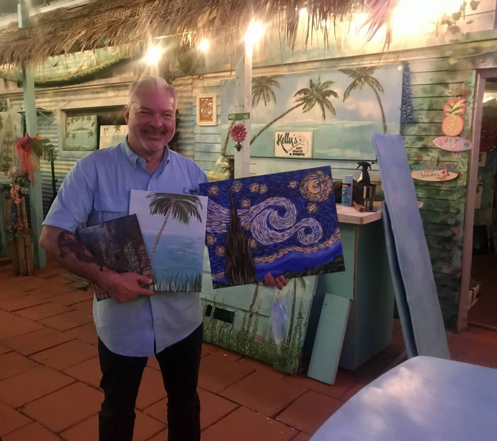 A man is holding two paintings in front of a thatched hut.