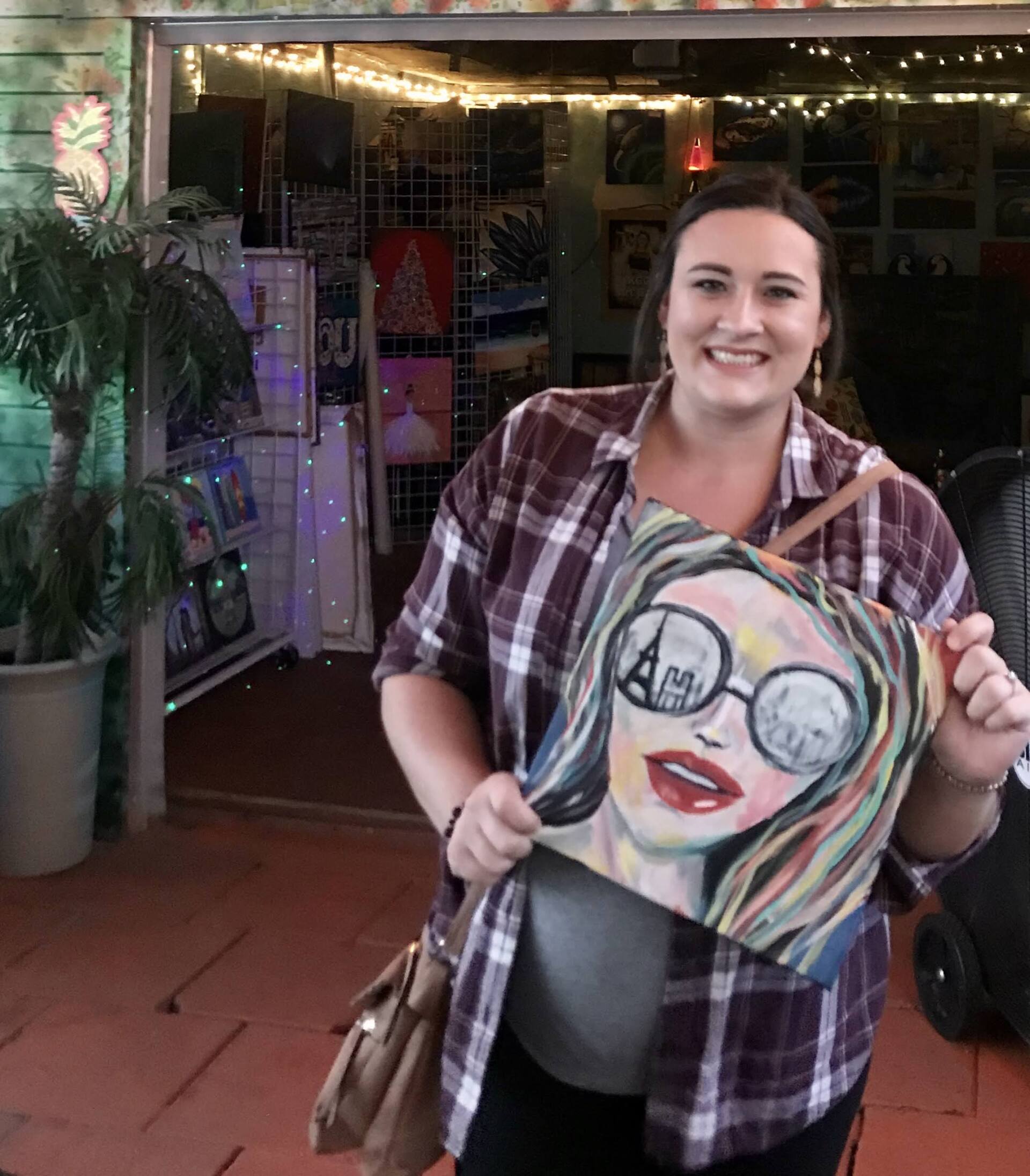 A woman in a plaid shirt is holding a painting of a woman wearing sunglasses