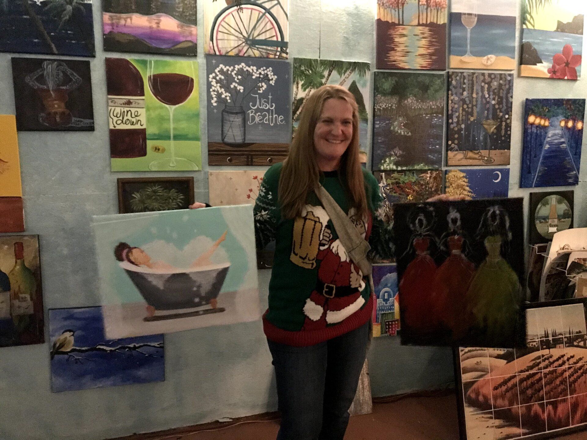 A woman in a santa sweater is standing in front of a wall of paintings.