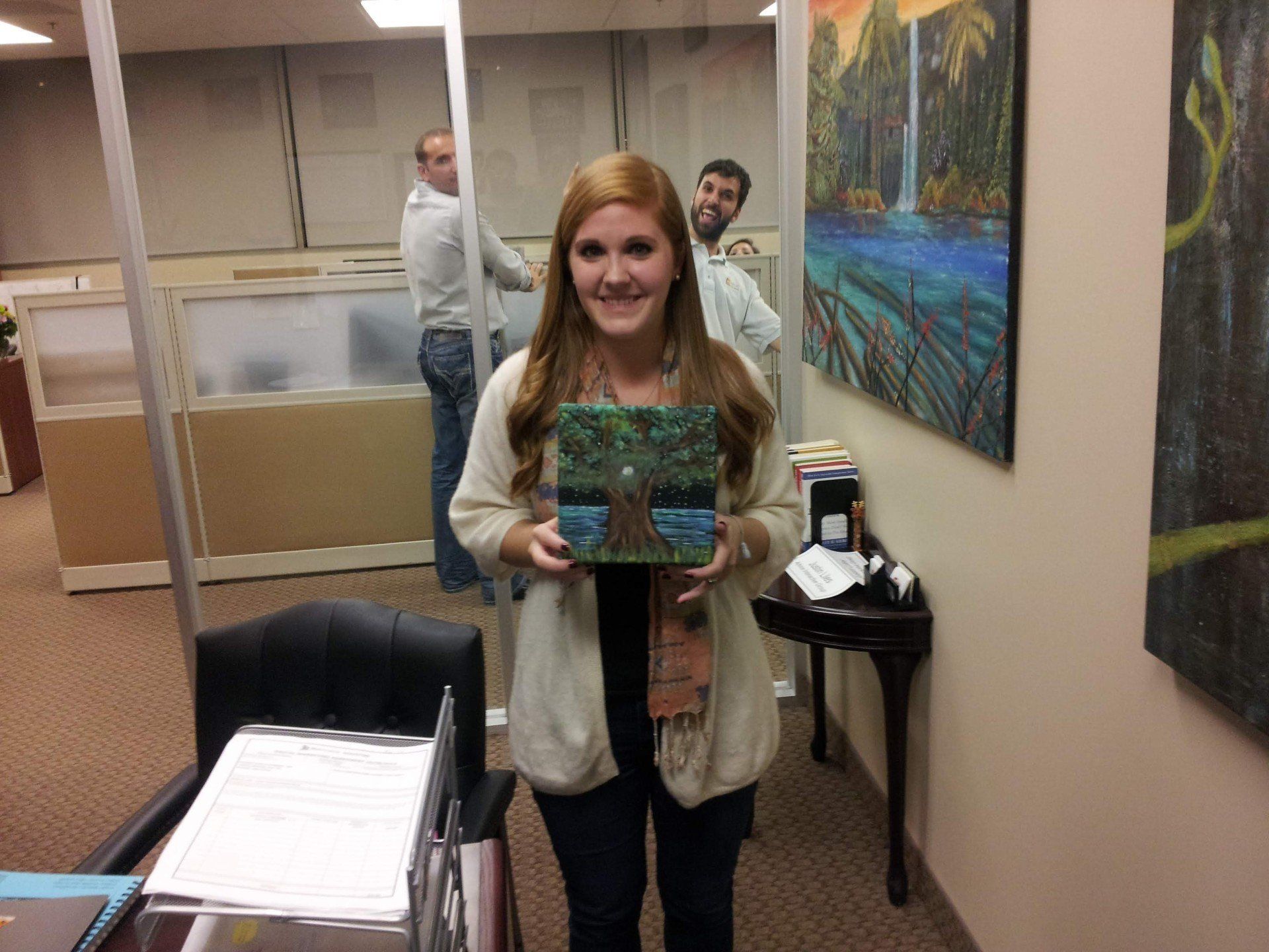 A woman is holding a painting of a tree in an office