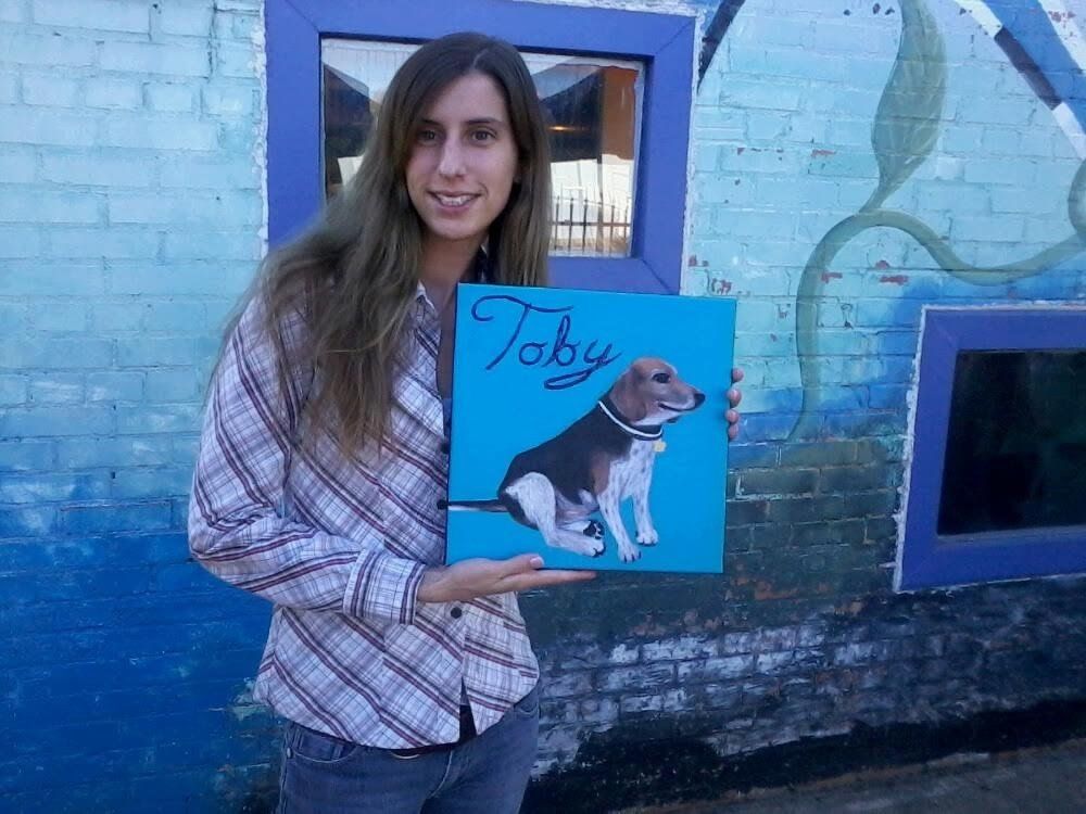 A woman is holding a painting of a dog named toby