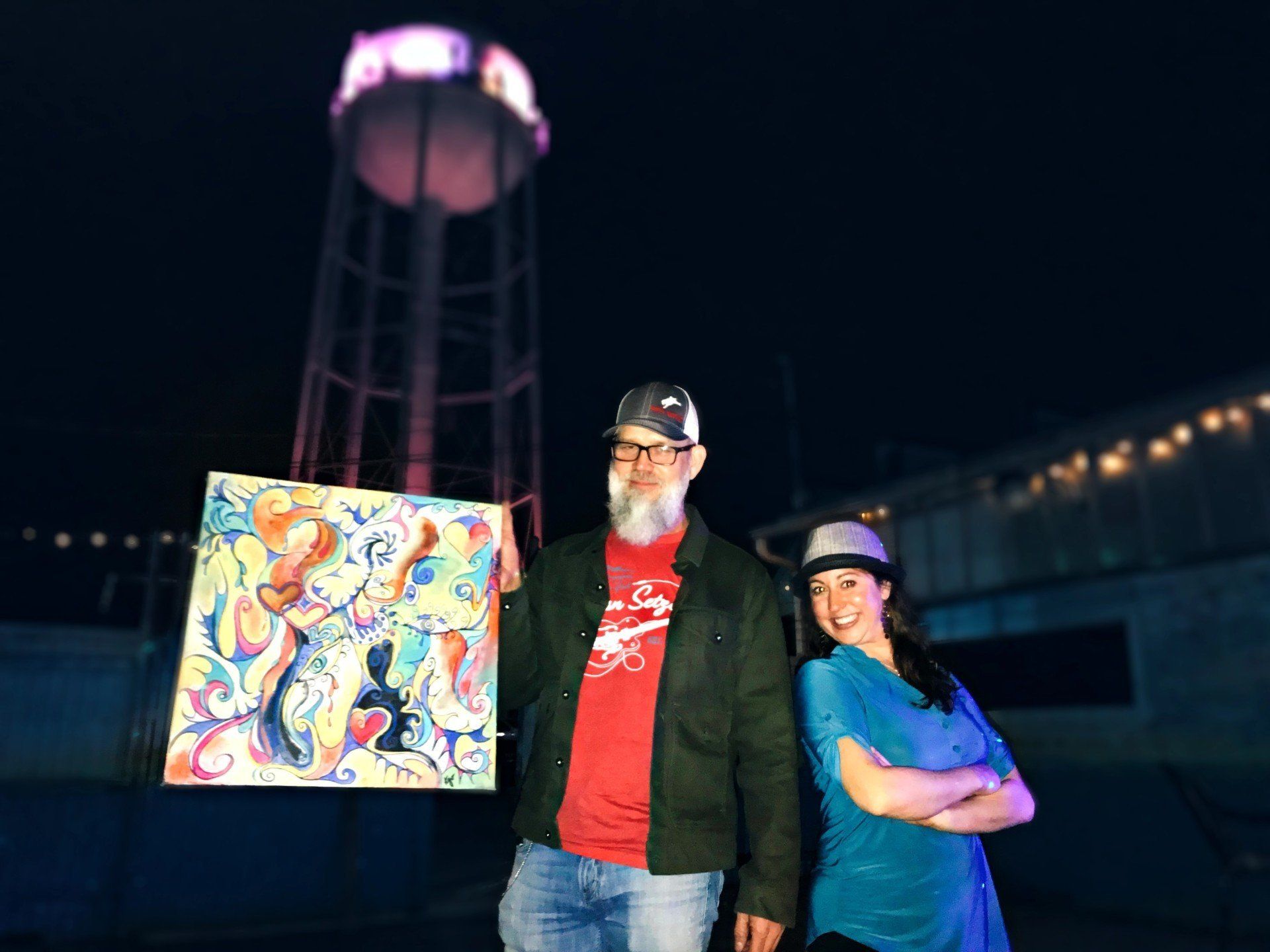 A man and a woman are standing next to each other holding a painting in front of a water tower.