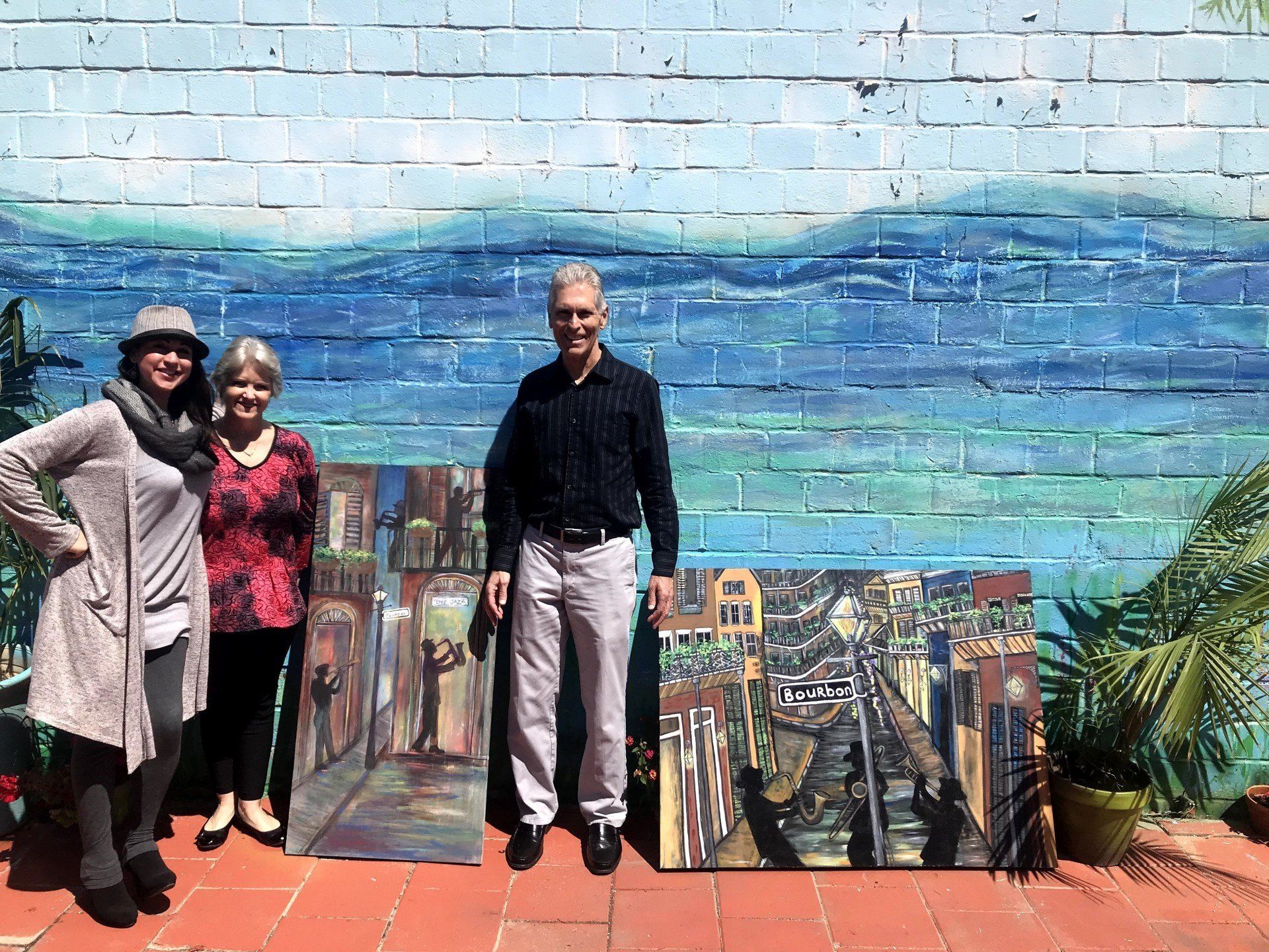 A group of people are standing in front of a brick wall holding paintings.