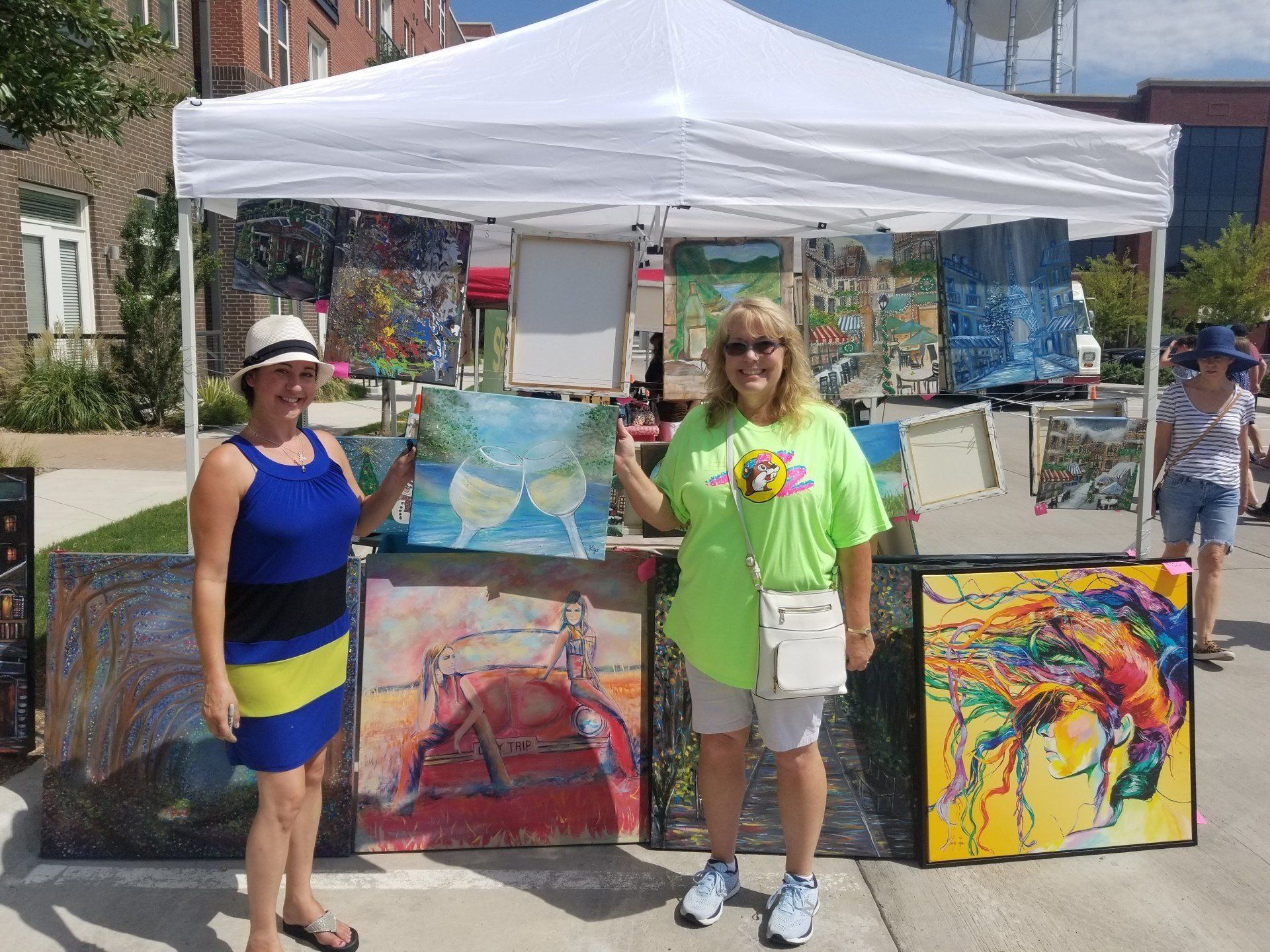 Two women are standing in front of a tent holding paintings.