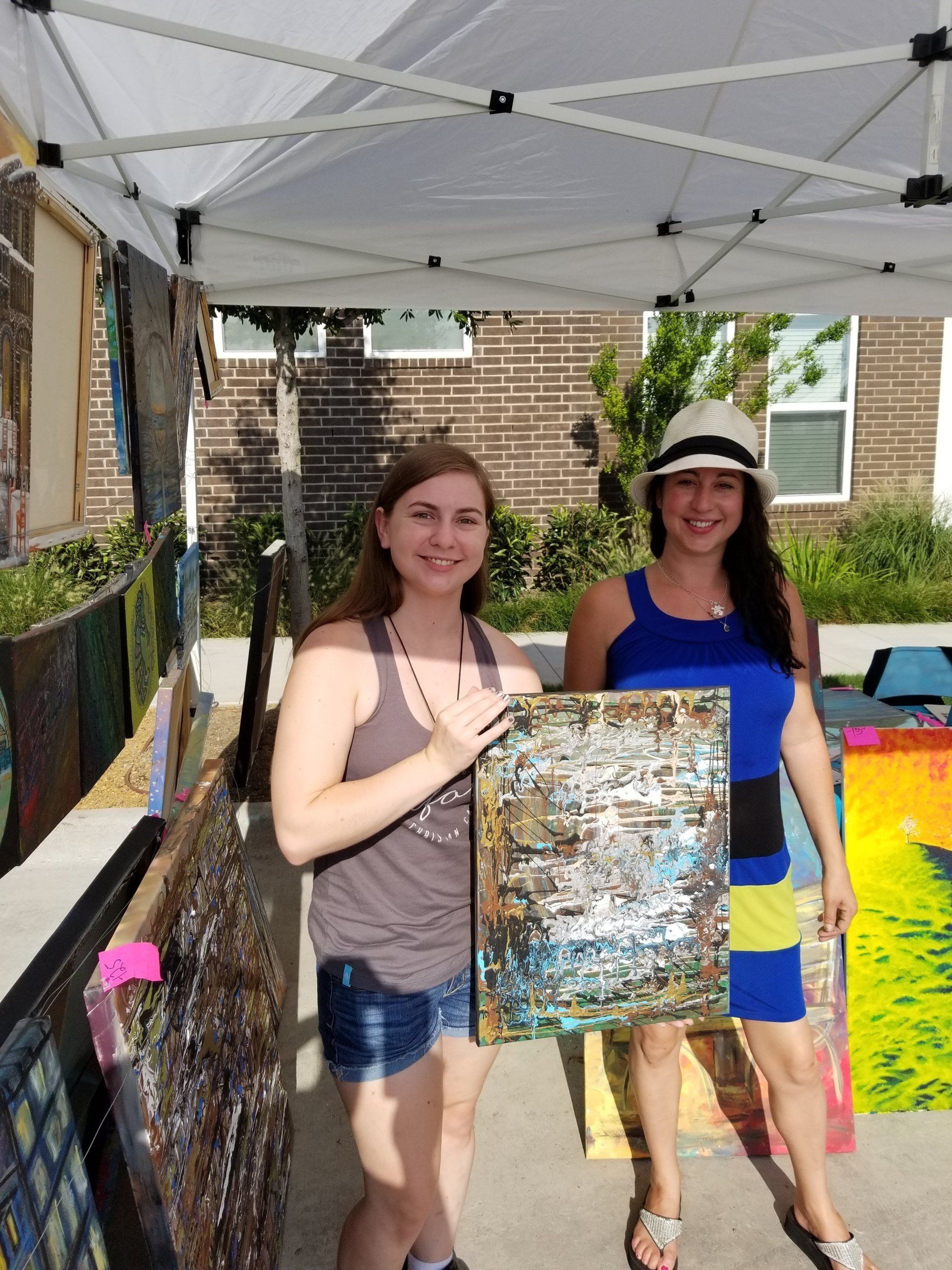 Two women standing under a tent holding a painting