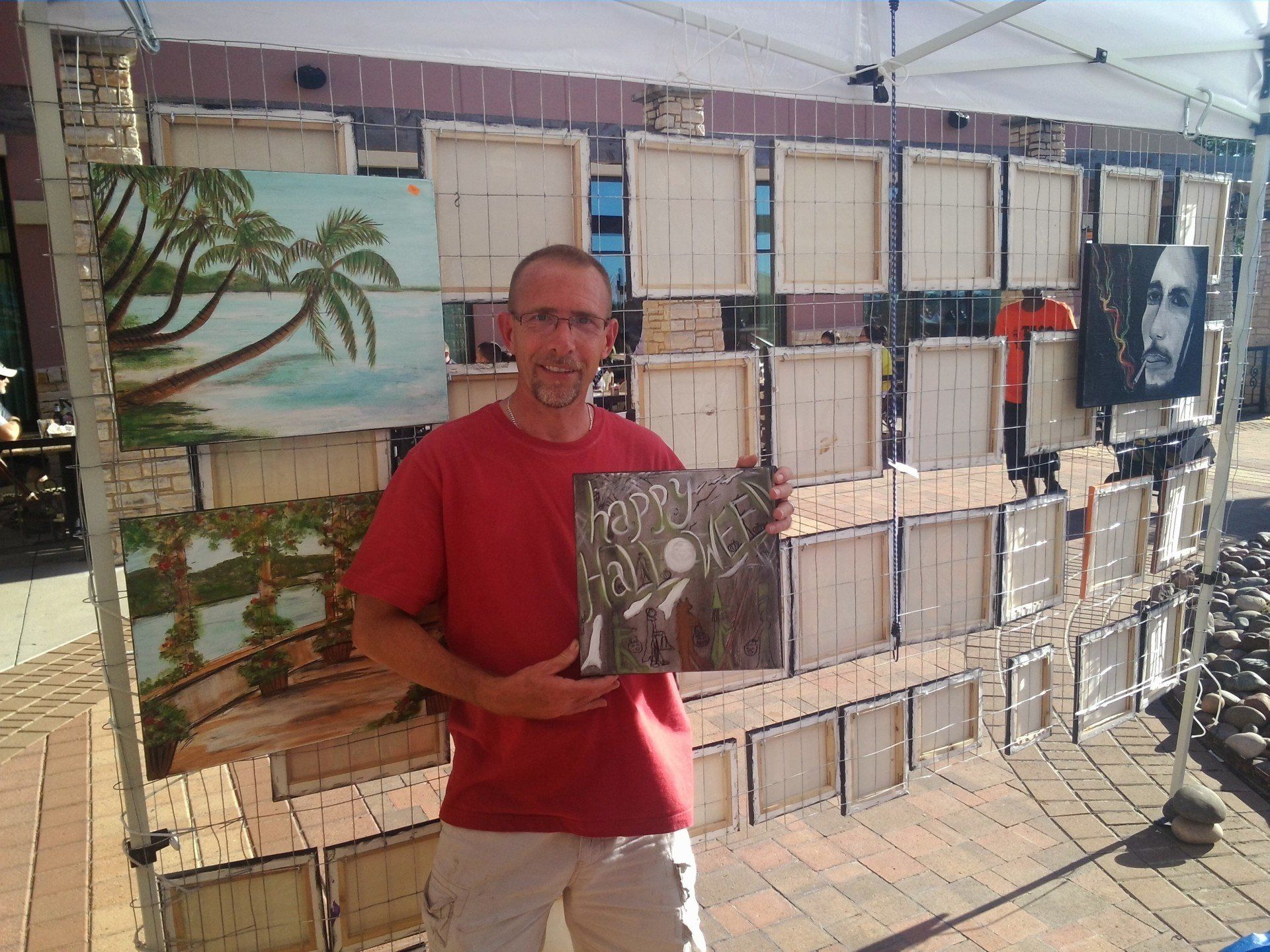 A man in a red shirt is holding a painting in front of a wall of paintings