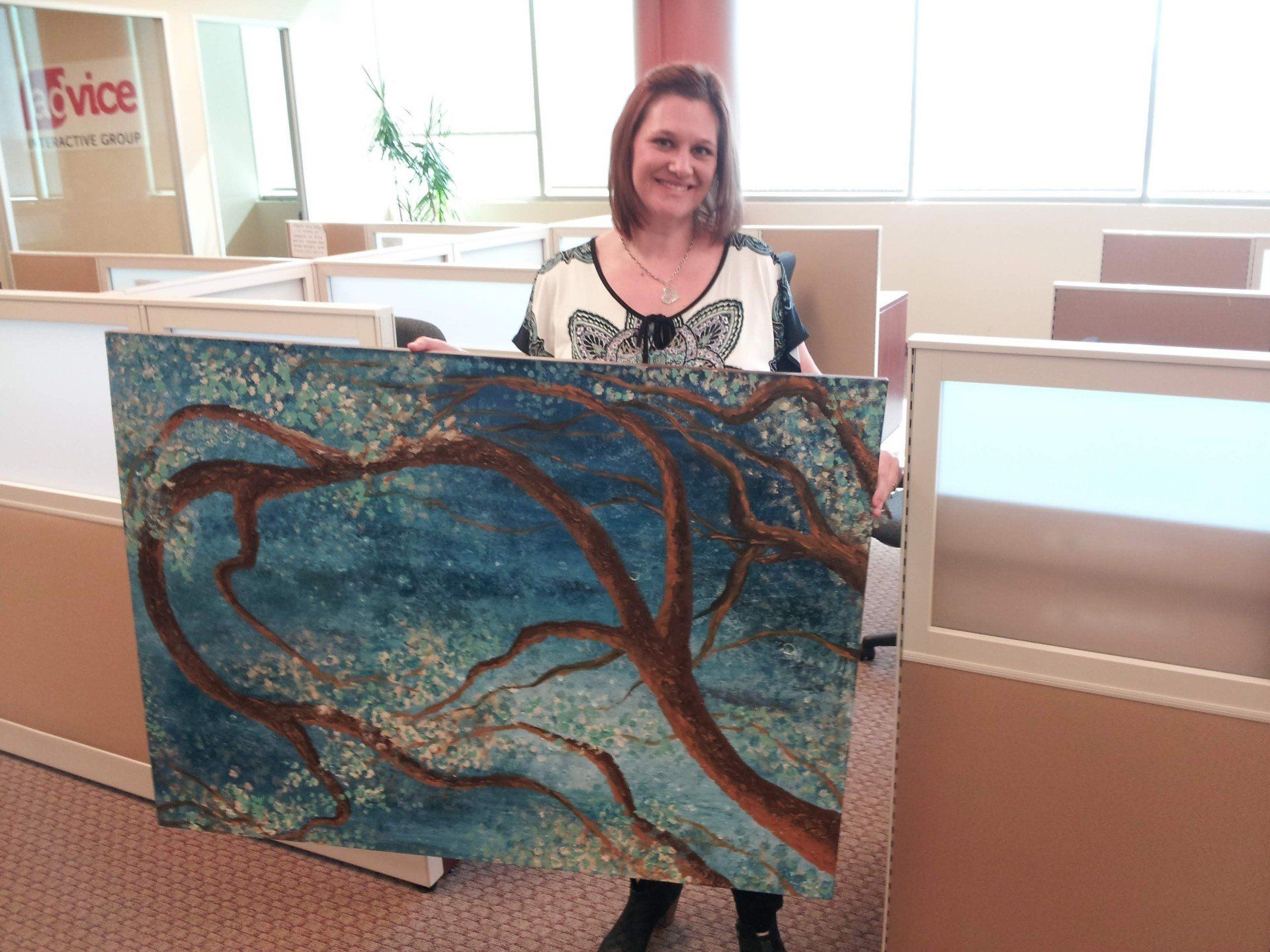 A woman is holding a large painting in an office cubicle.