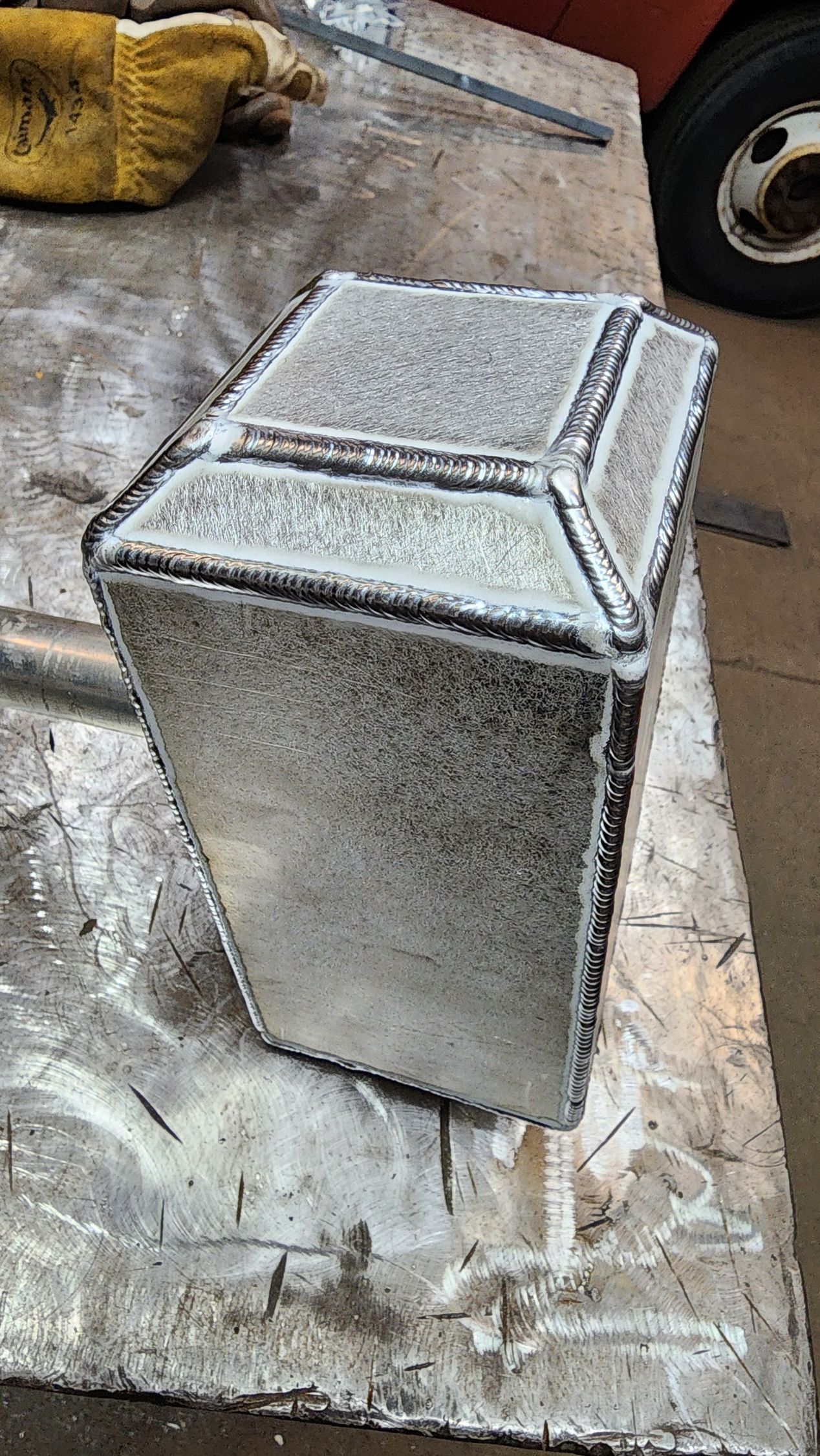 Welded metal box on a workbench, bright silver with dark welds, in a workshop setting.