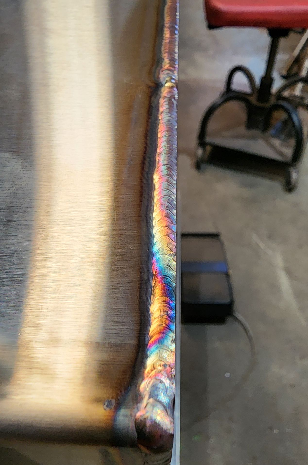 Close-up of welded metal, showing colorful heat marks along the seam. In a workshop setting, near a stool.