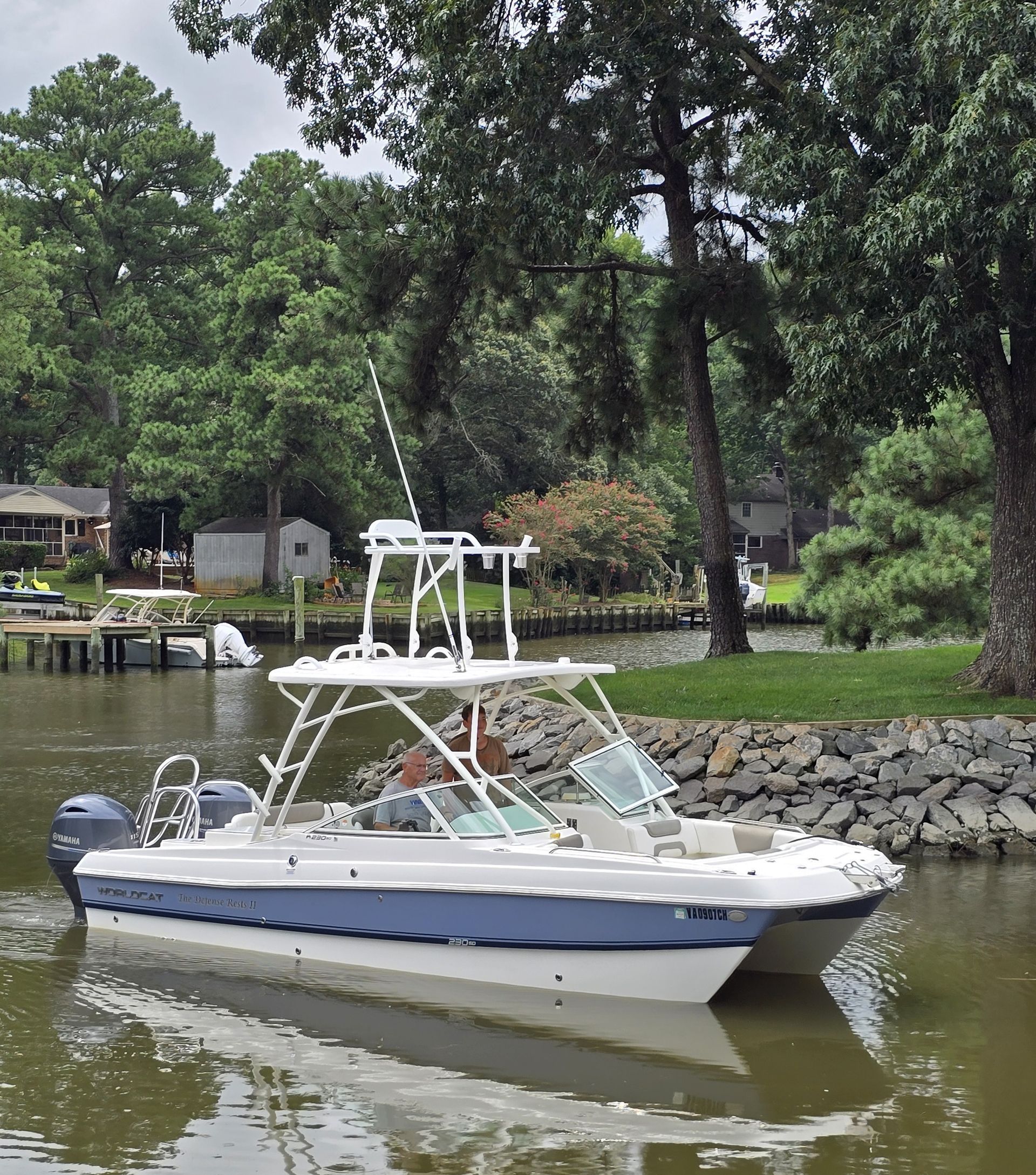 White and blue catamaran boat on water, two people visible. Trees and houses in background.