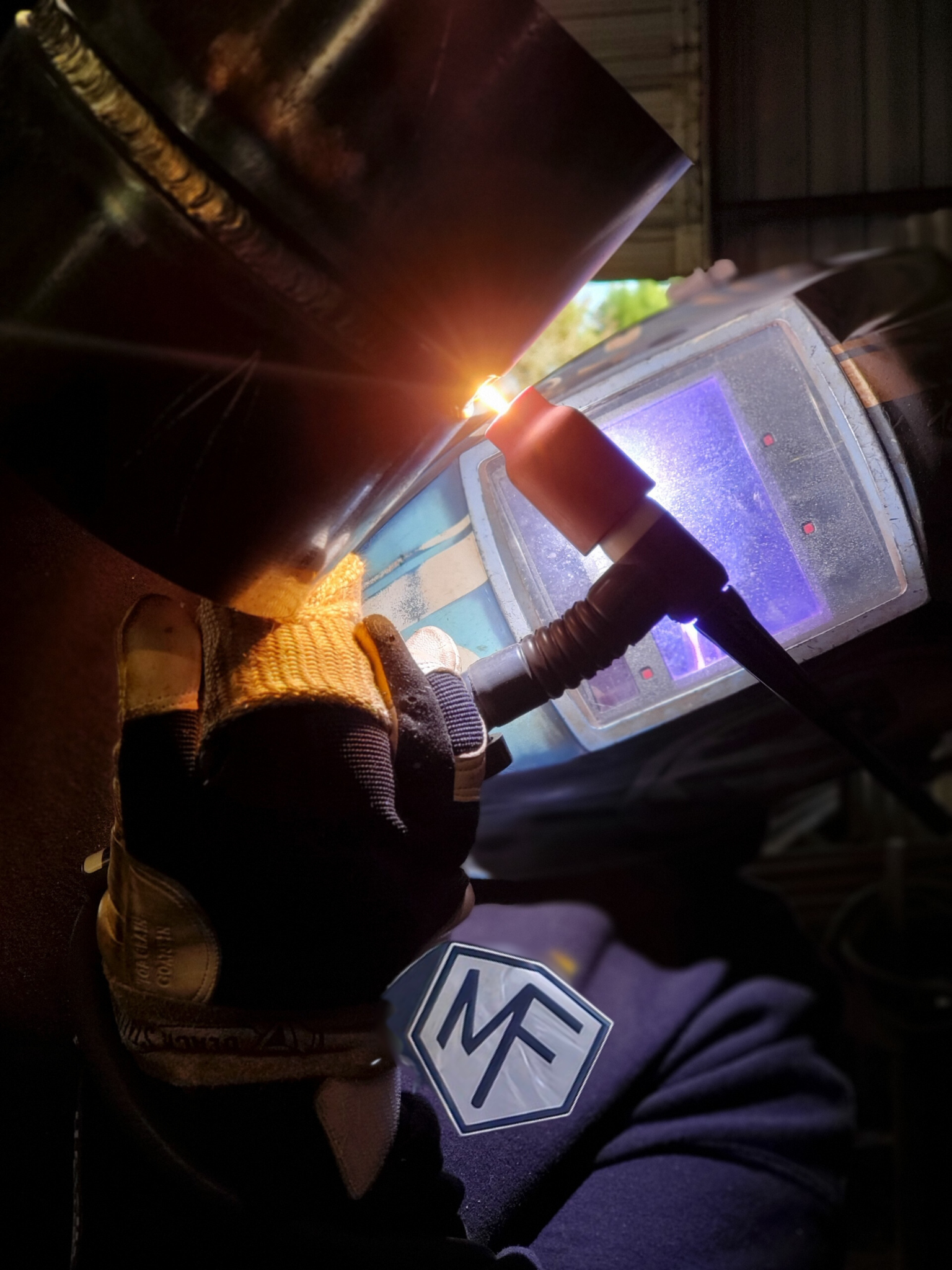 Welder in protective gear welding metal pipes, with bright arc, indoor setting.