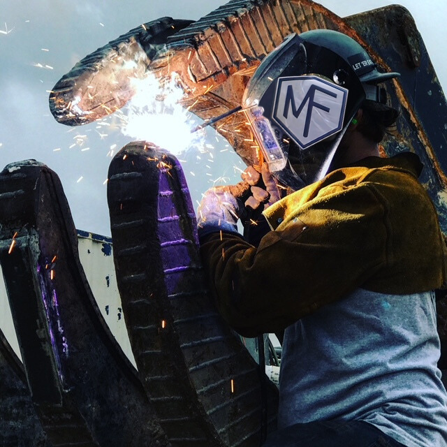 Welder in action; welding machinery with sparks flying. Wearing a helmet, gloves and jacket outdoors.