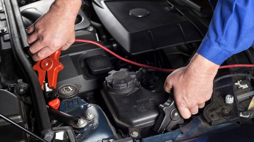 Mechanic connecting jumper cables to a car battery in an engine bay; red and black cables.