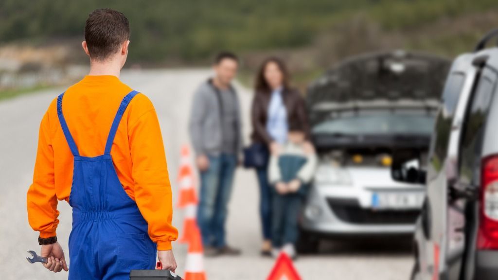 Mechanic in orange and blue jumpsuit assisting a family with a broken-down car on a roadside.