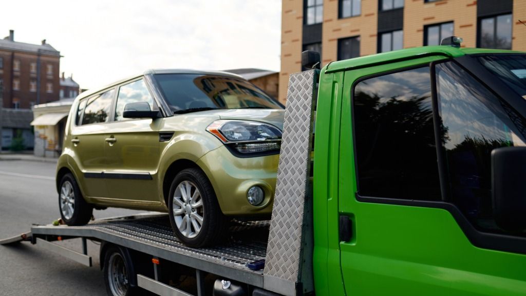 A gold Kia Soul is being towed by a green tow truck on a city street.