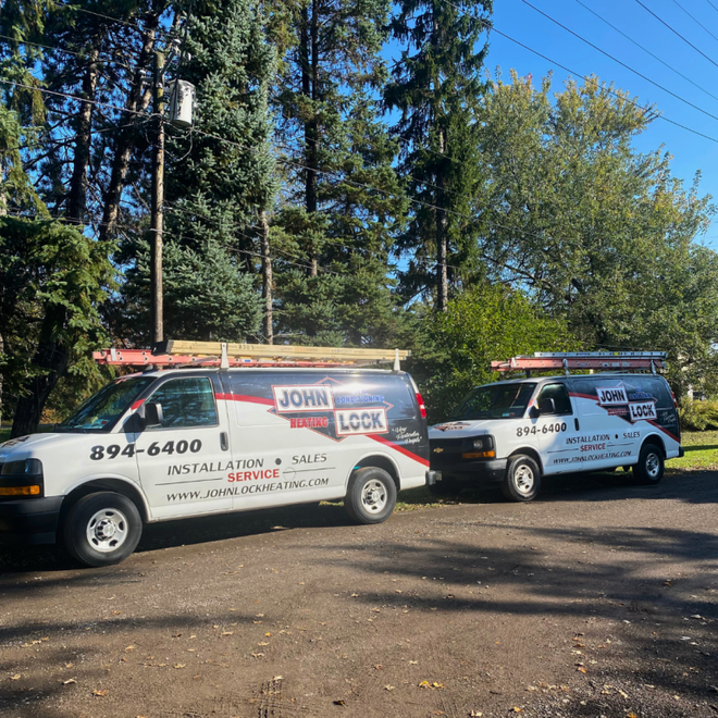 Two white work vans for John Lock Plumbing & Heating parked on a driveway in front of a tree line on a sunny day.