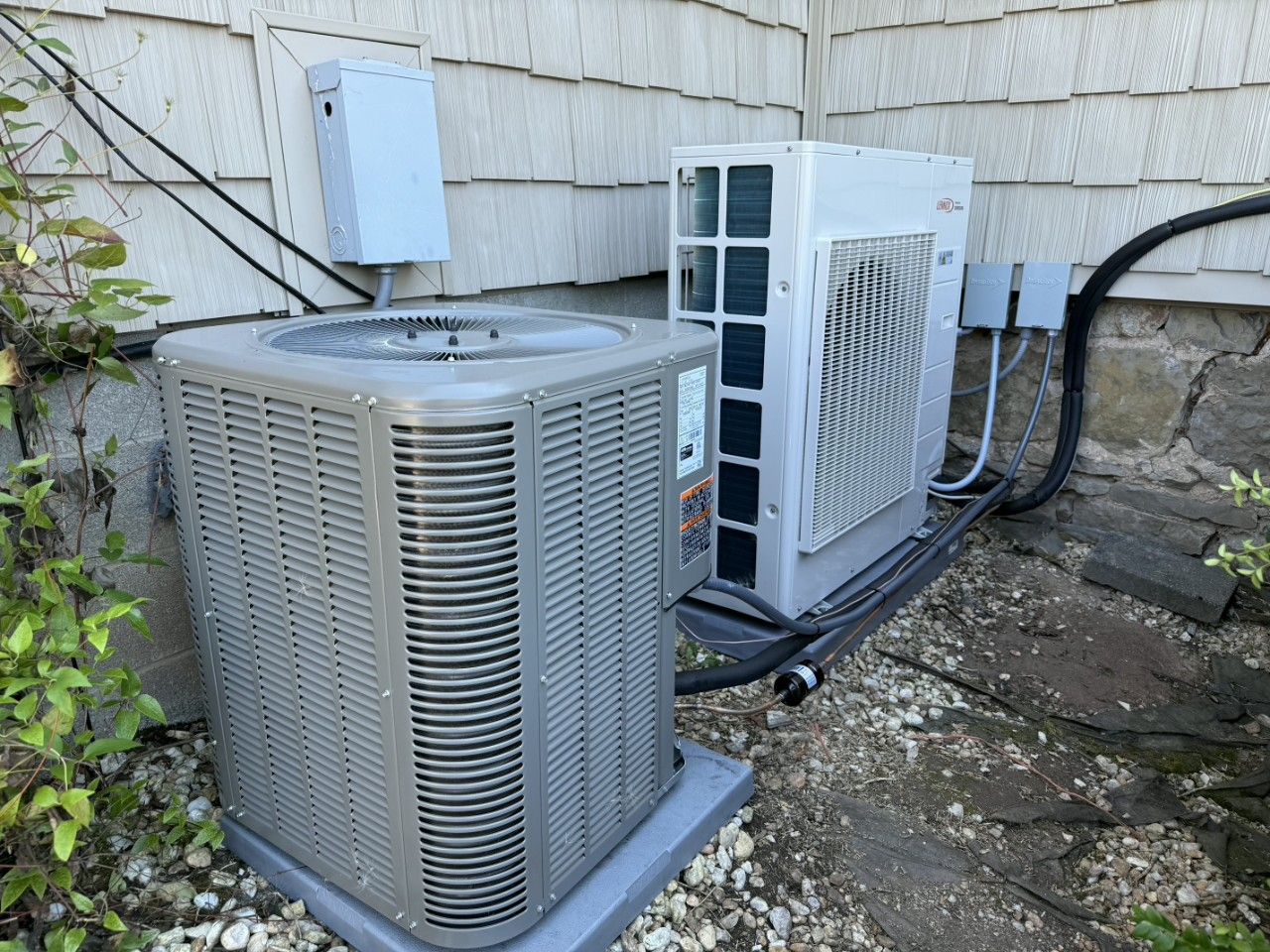 Two outdoor HVAC units sitting on a gravel surface against a house wall with cedar shake siding.