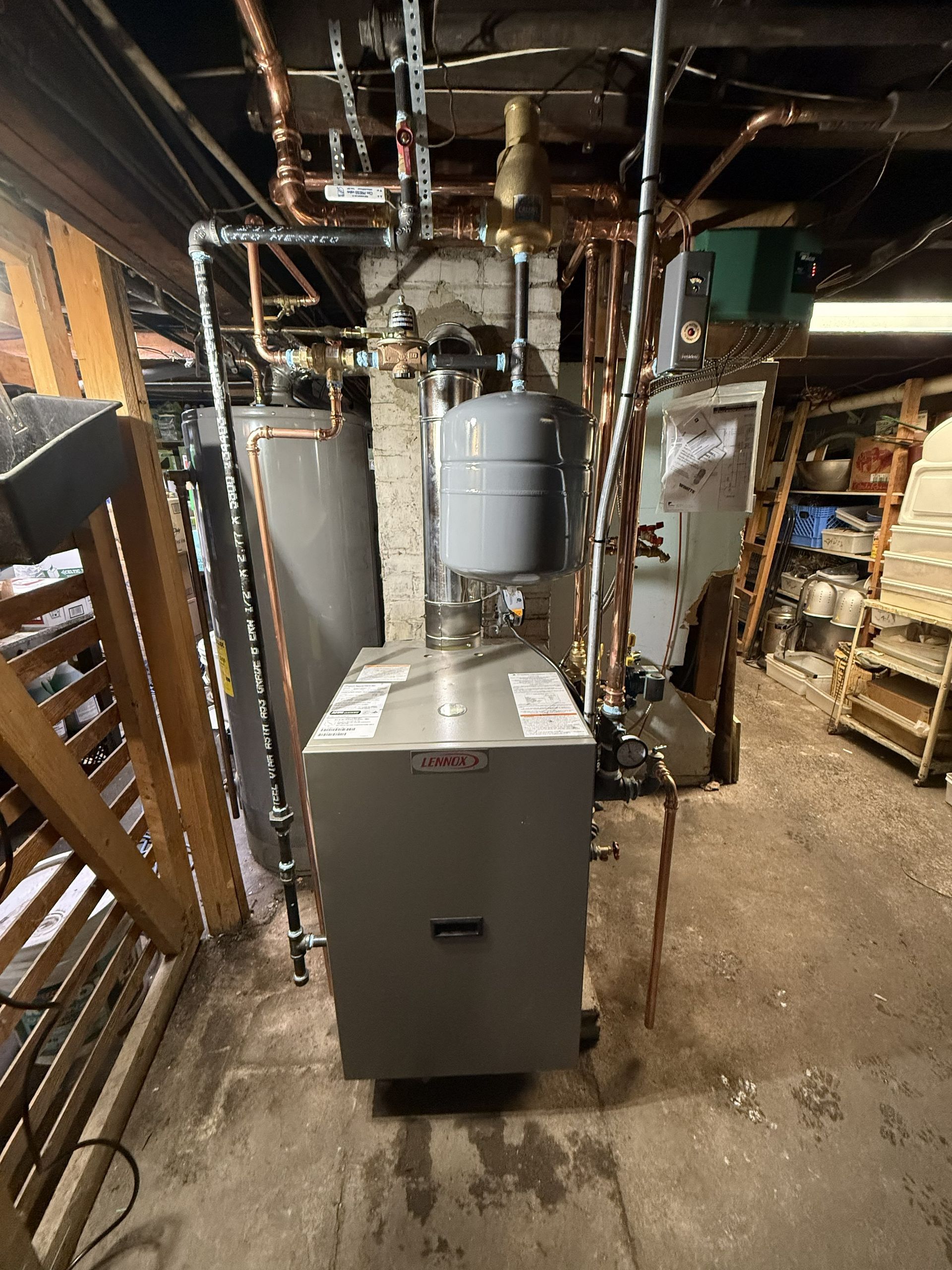 A grey residential boiler system with connected copper piping, an expansion tank, and a vent pipe in a basement setting.