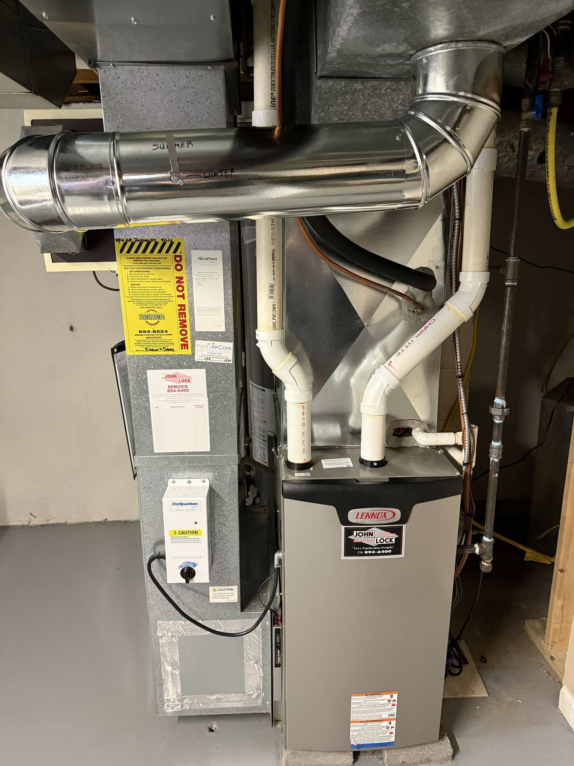 A vertical Lennox furnace unit in a basement, connected to a silver metal duct and two white PVC exhaust pipes.