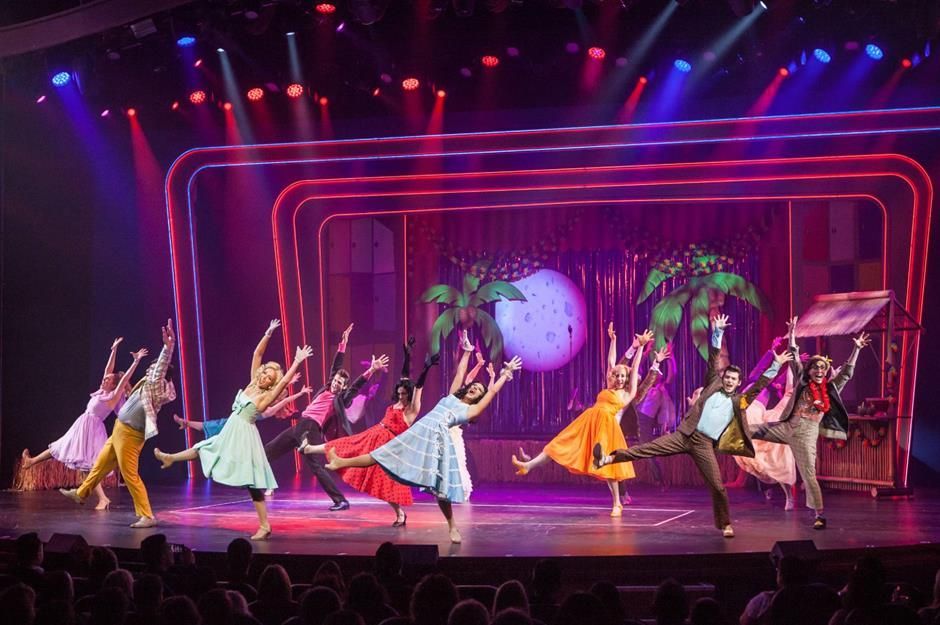 Stage performers dancing in colorful costumes, neon lights, and a backdrop of palm trees and a moon.