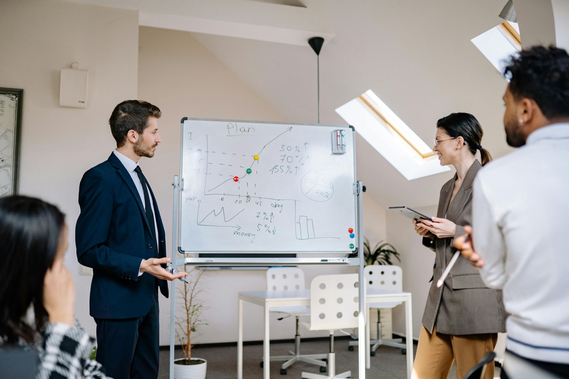 Business team in a meeting; man presents a whiteboard, others observe and hold devices in a bright office.