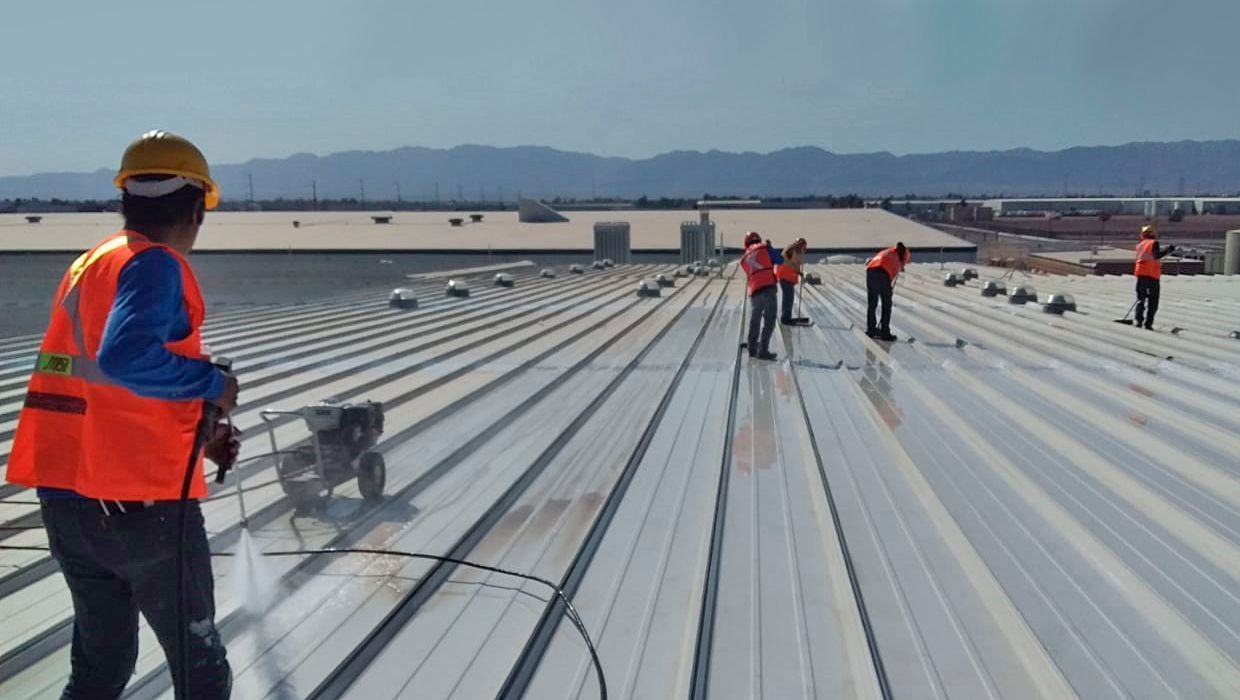 Trabajadores con chalecos reflectantes utilizan equipos de pulverización para limpiar un gran techo metálico industrial bajo un cielo brillante.