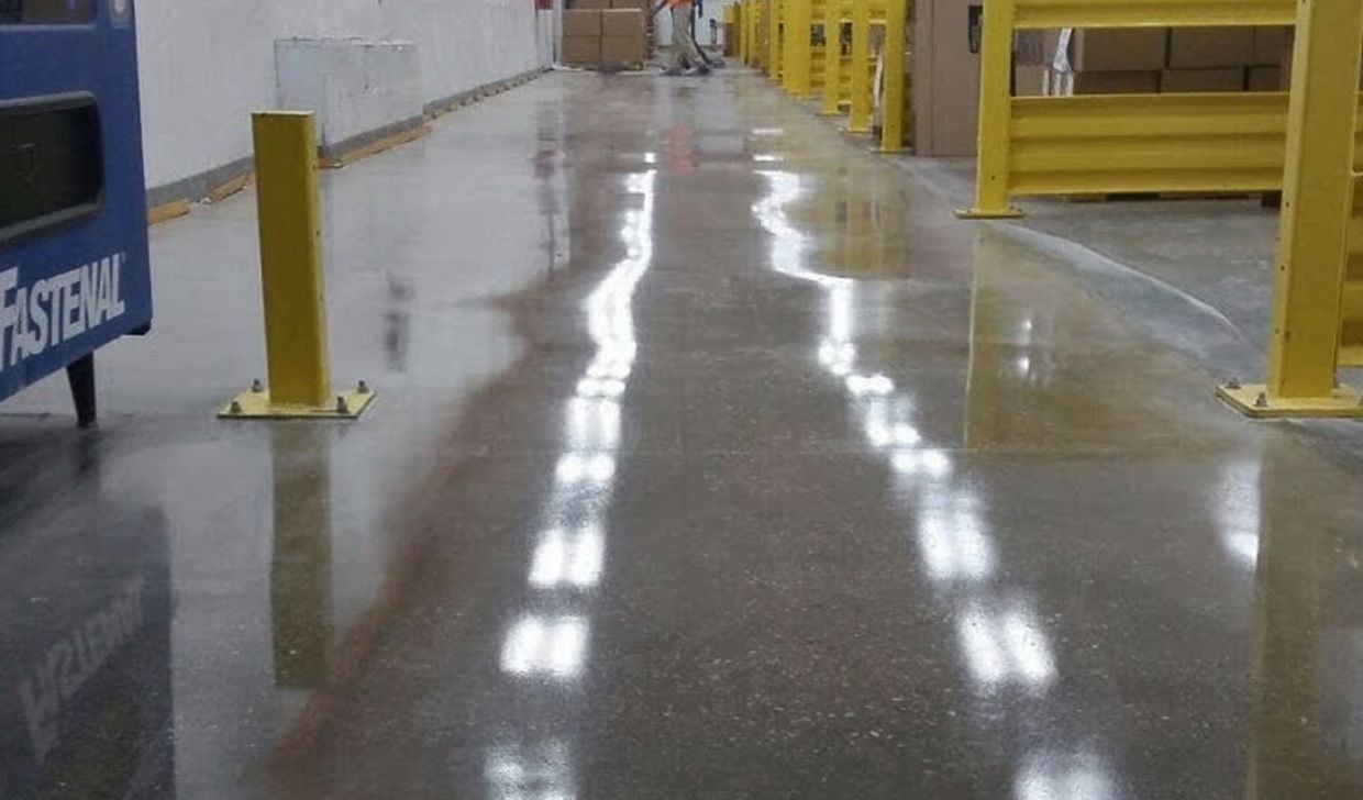 El suelo de hormigón pulido de un almacén refleja las luces del techo, dejando ver los bolardos de seguridad amarillos y las estanterías.