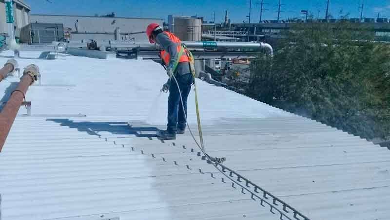 Un trabajador con chaleco reflectante y casco utiliza una pistola pulverizadora para recubrir un tejado de metal blanco.