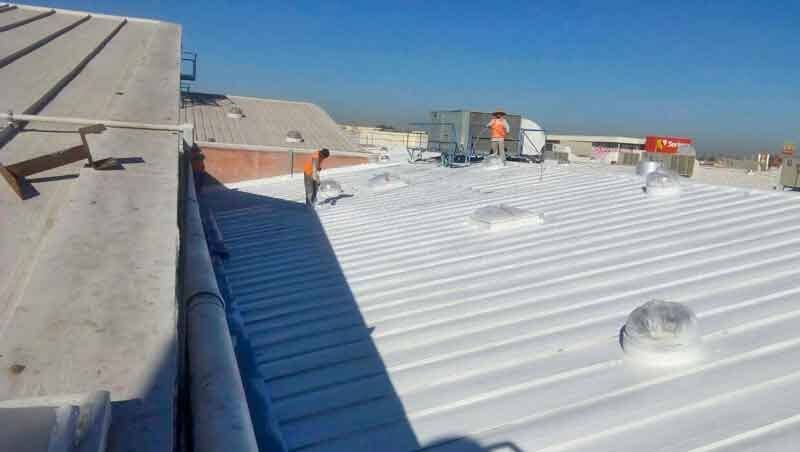 Dos trabajadores en un tejado de chapa ondulada blanca durante un proyecto de mantenimiento o recubrimiento, bajo un cielo azul despejado.