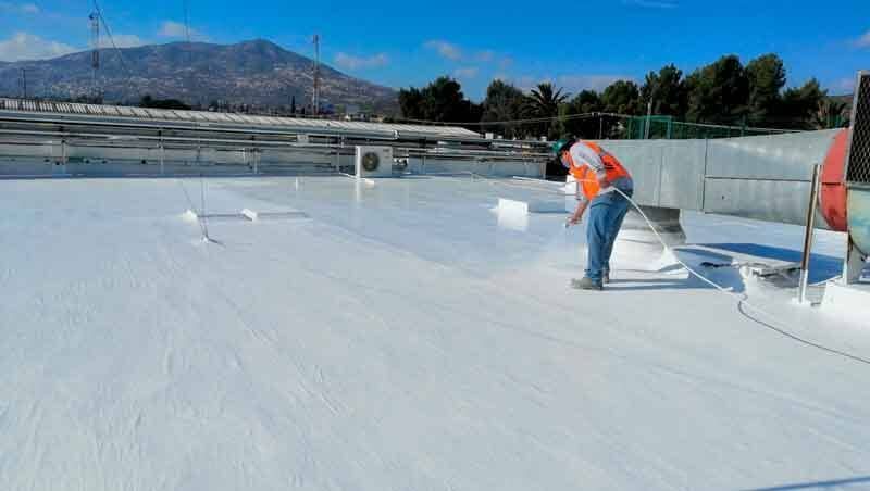 Un trabajador con un chaleco de seguridad naranja rocía una capa protectora blanca sobre un tejado plano al aire libre, bajo un cielo azul brillante.
