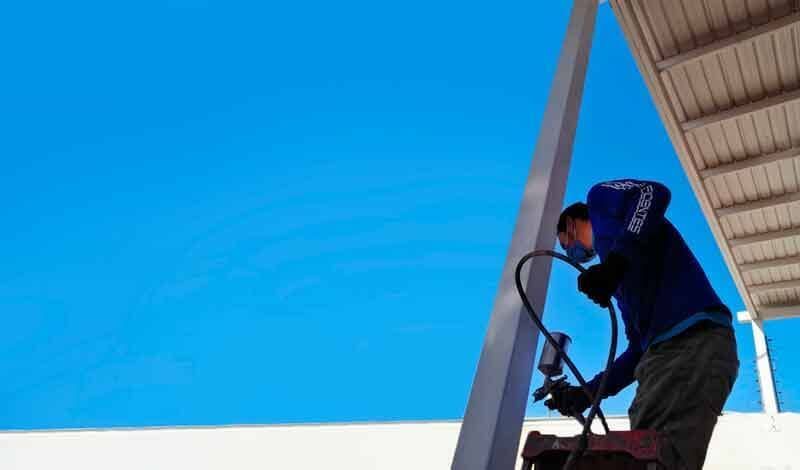Un trabajador con camisa azul utiliza una herramienta de pintura en aerosol para pintar una viga metálica vertical bajo un techo parcial, con el cielo azul de fondo.