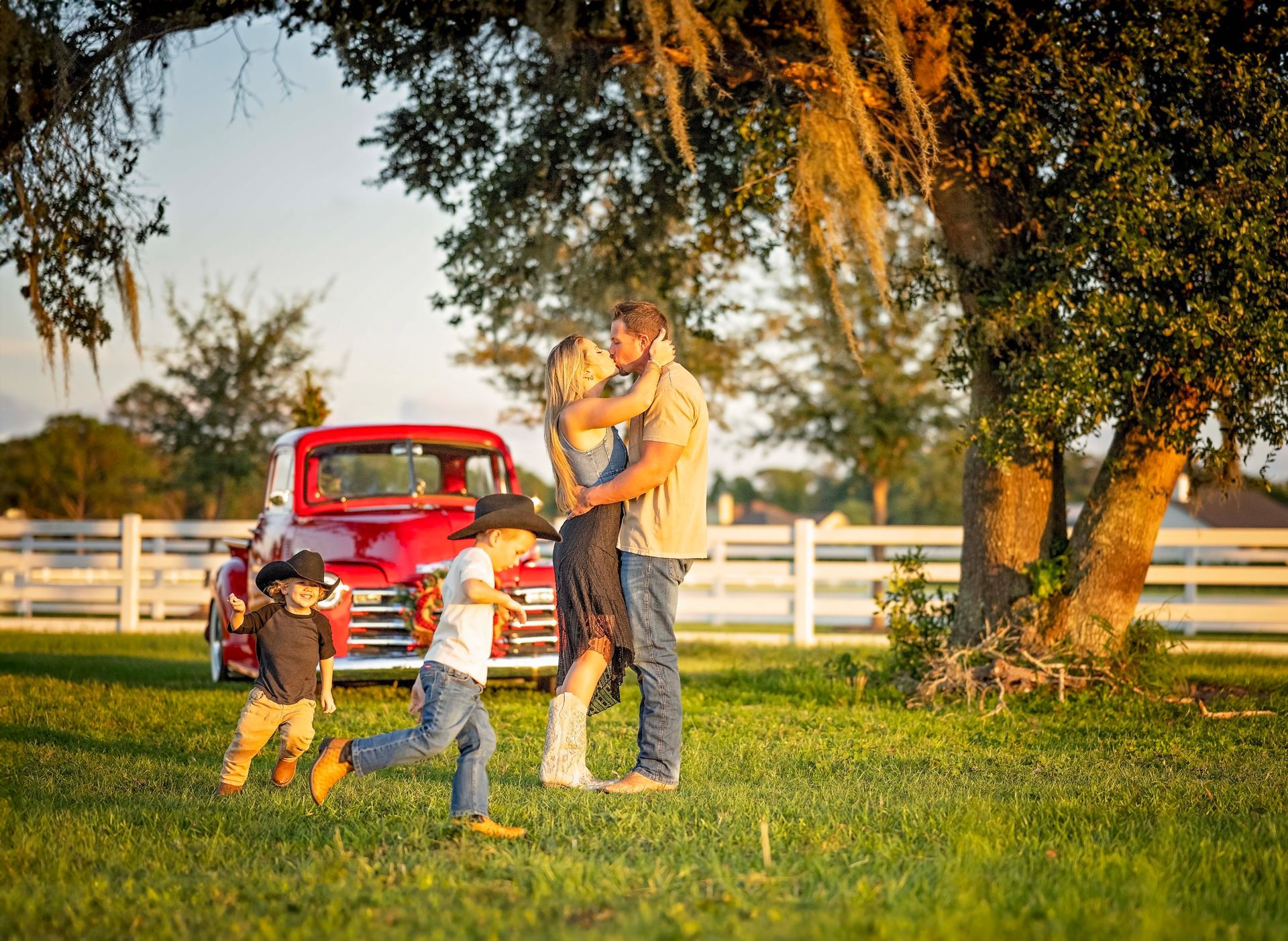 Family embracing in a field with a red truck, boys running nearby. Golden hour.