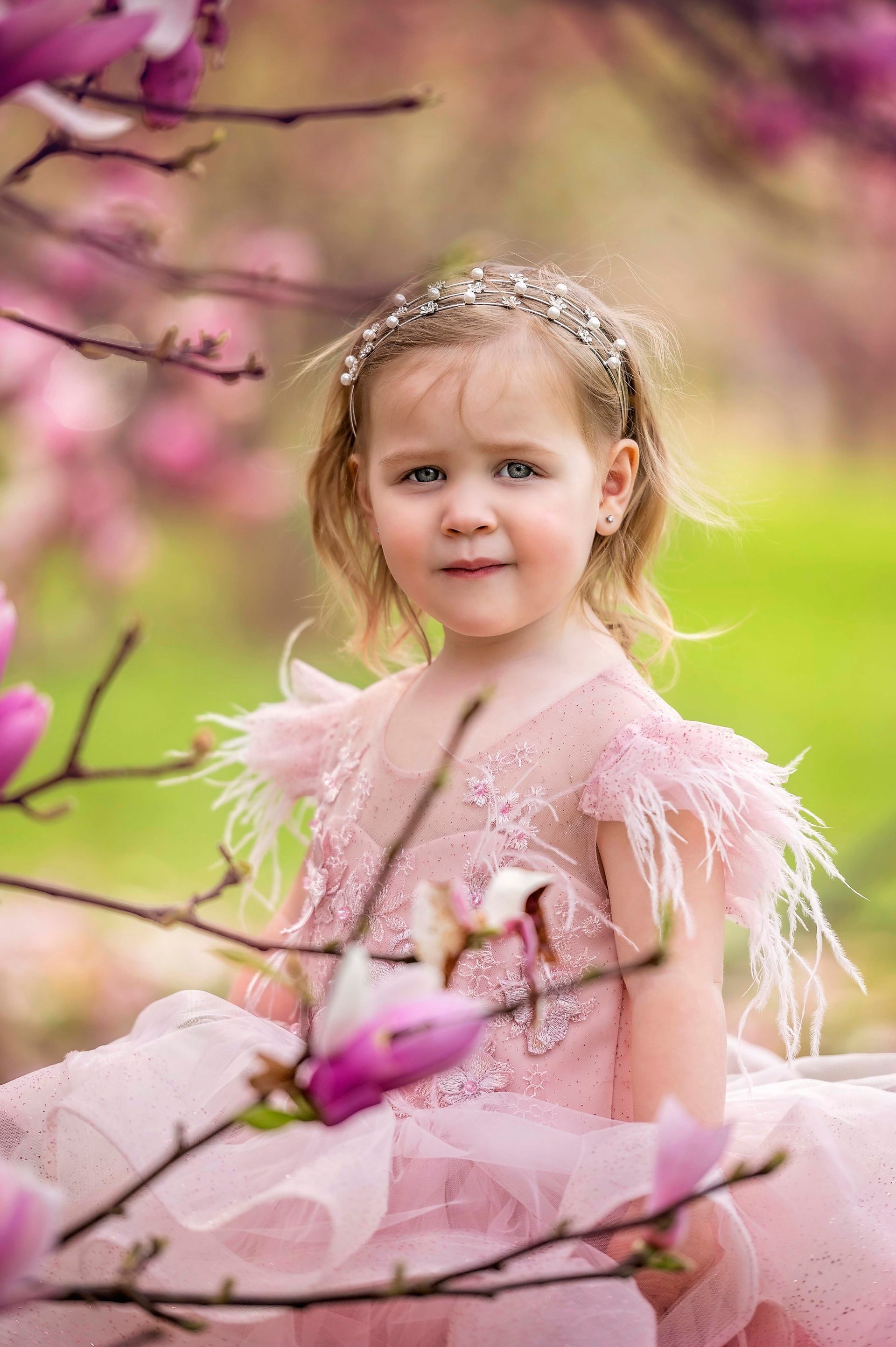 Young girl in pink dress and bejeweled headband poses among pink flowers.