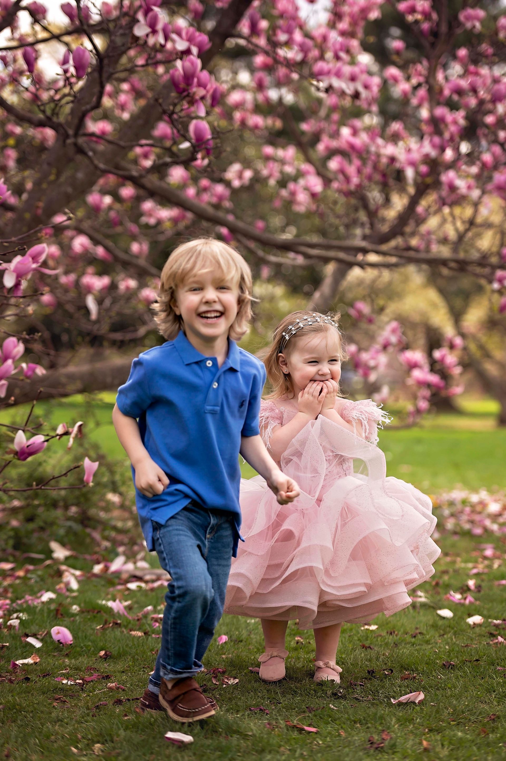 Boy in blue shirt and girl in pink dress laugh and run under a blooming pink tree in a park.