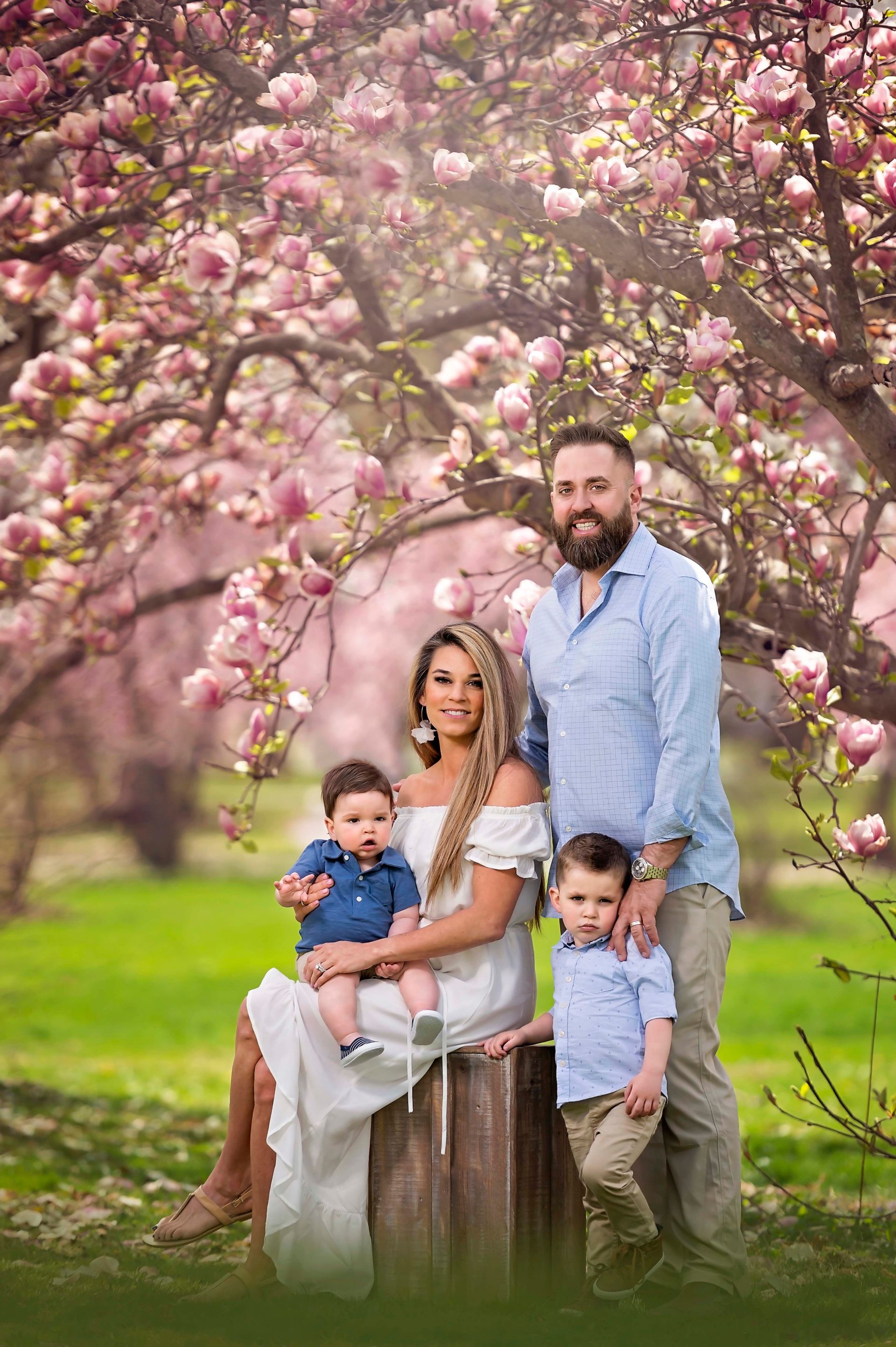 Family of four poses under blooming pink tree in a park. Mother holds baby; father stands next to older child.