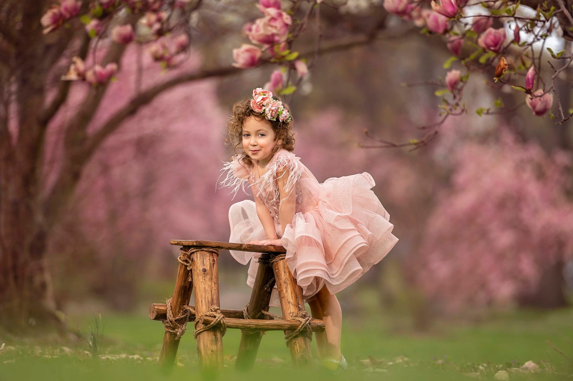 Girl in pink dress stands on wooden stool under flowering tree.