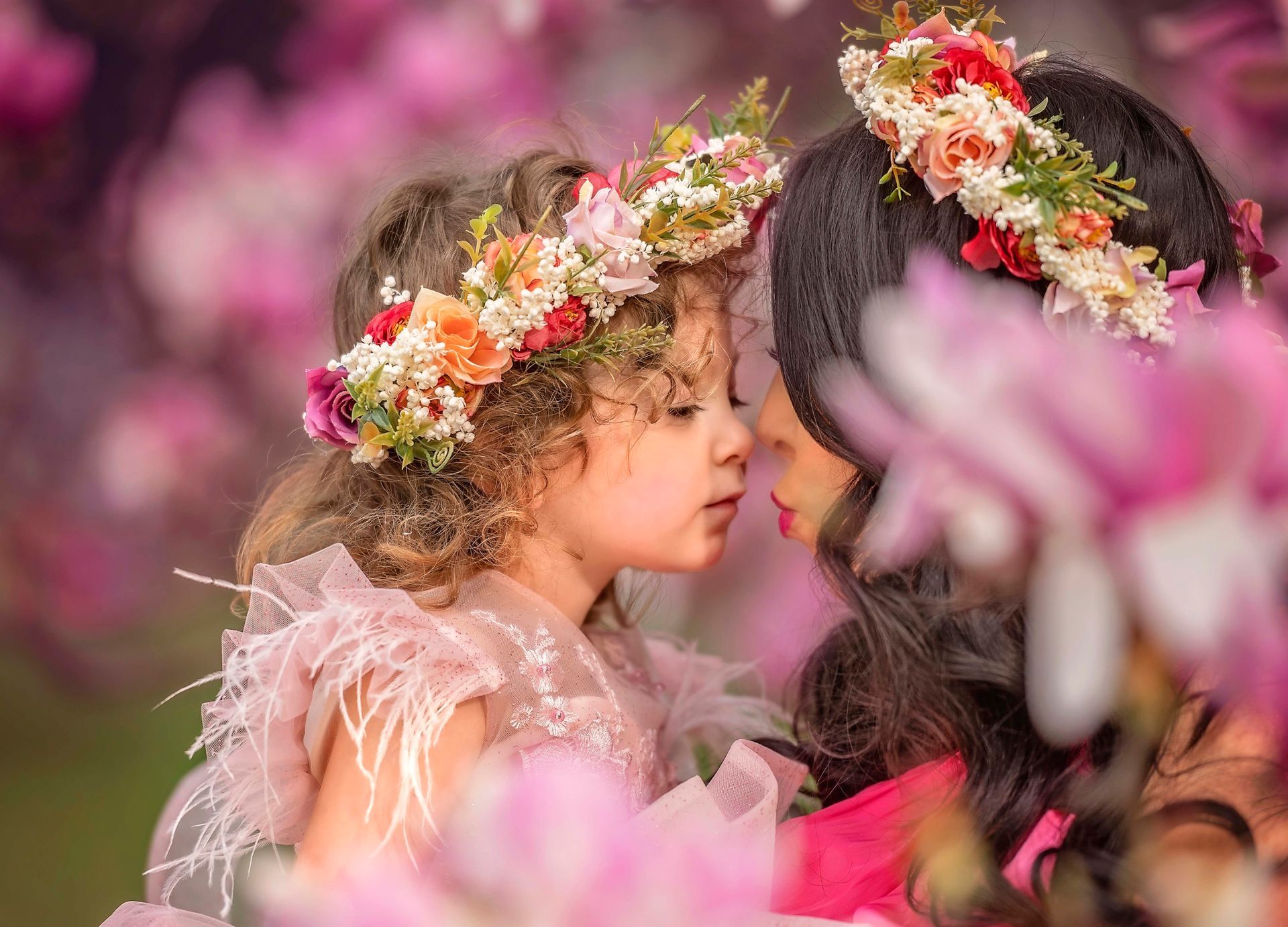 Mother and daughter in flower crowns, touching noses, surrounded by pink blossoms.