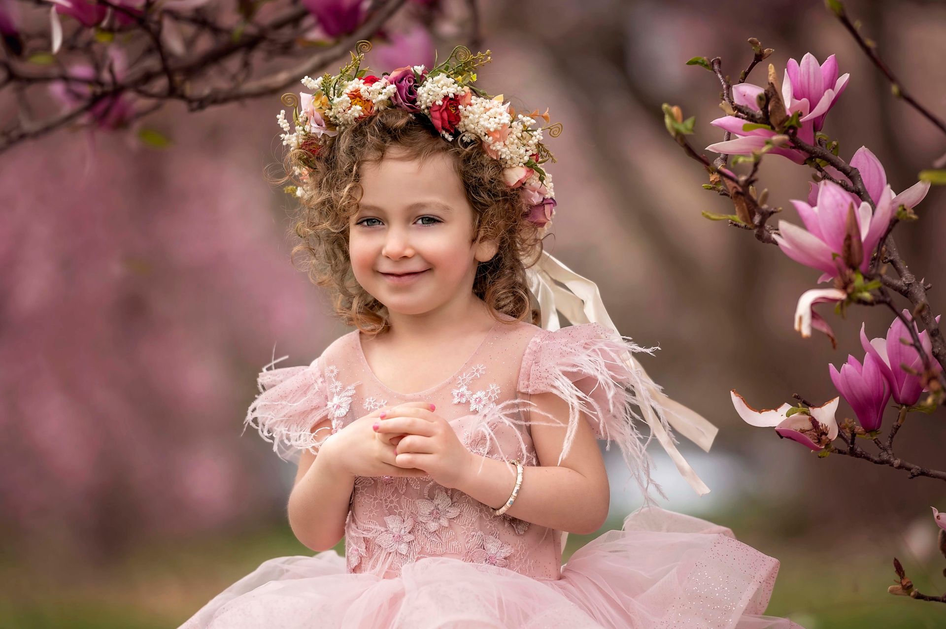 Young girl in pink dress and floral crown smiles outdoors among pink flowers.