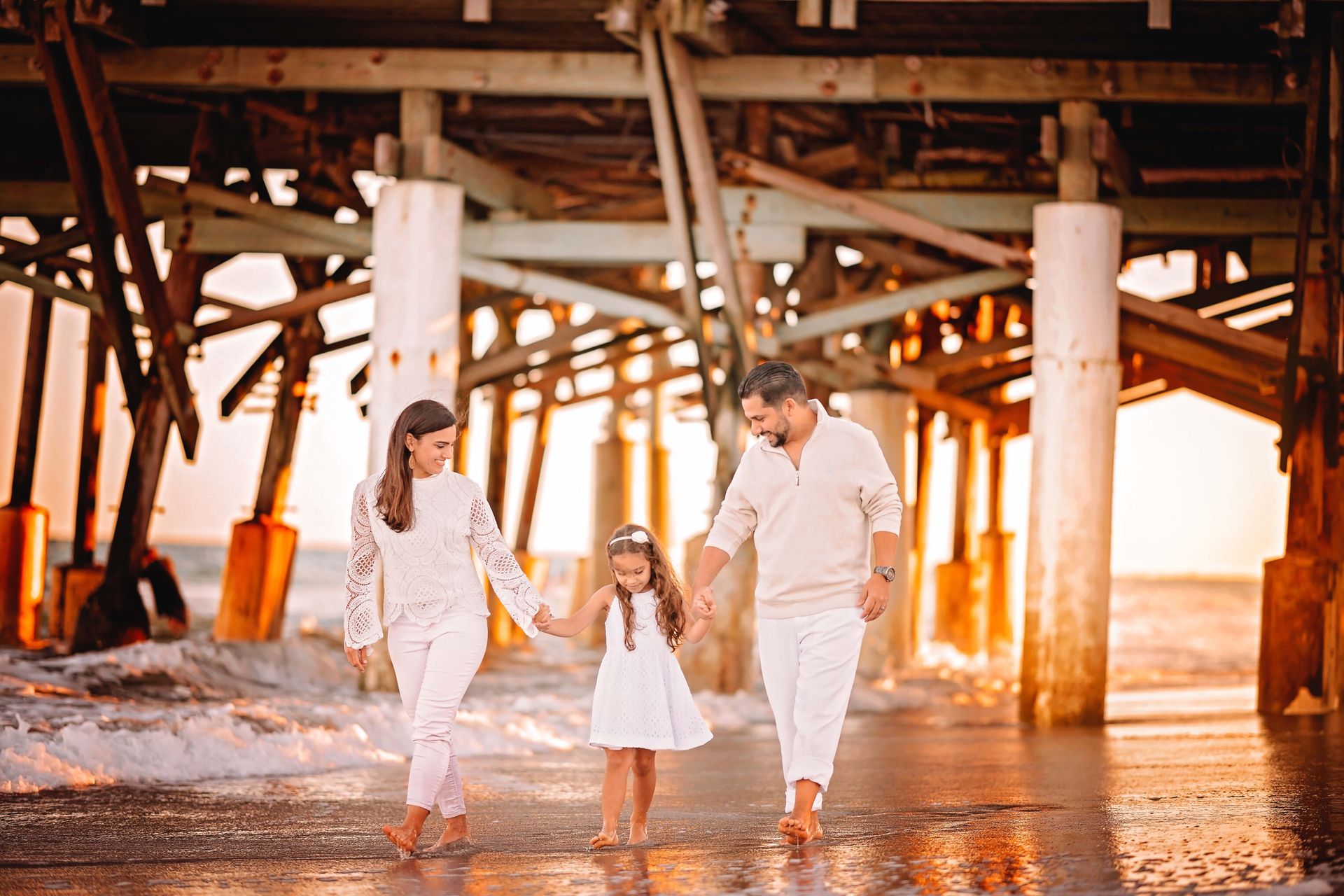 Family of three walking on beach under a pier, holding hands. They are all wearing white clothing.