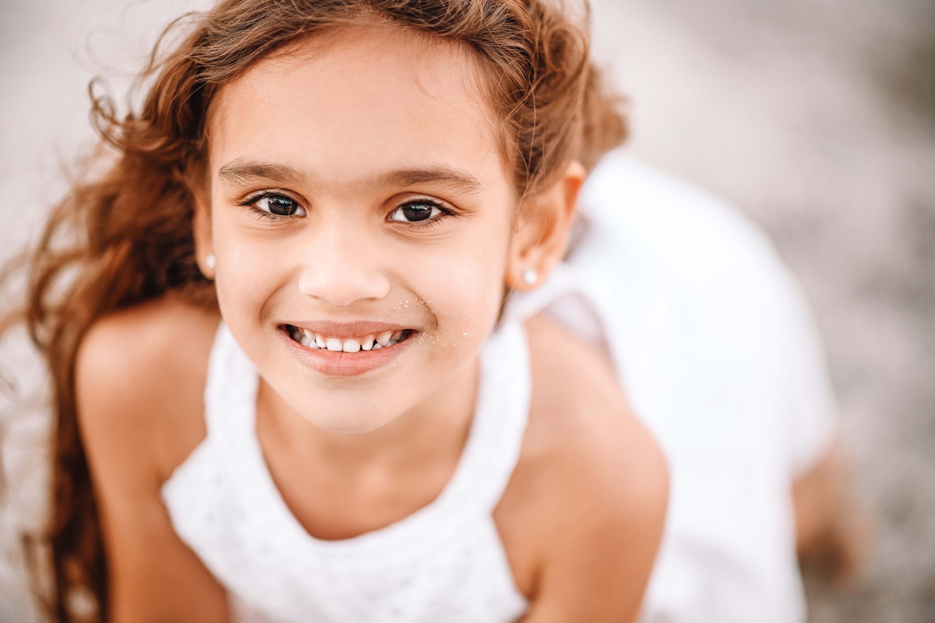 Smiling girl with long brown hair wearing a white dress, outdoors on a sandy beach.
