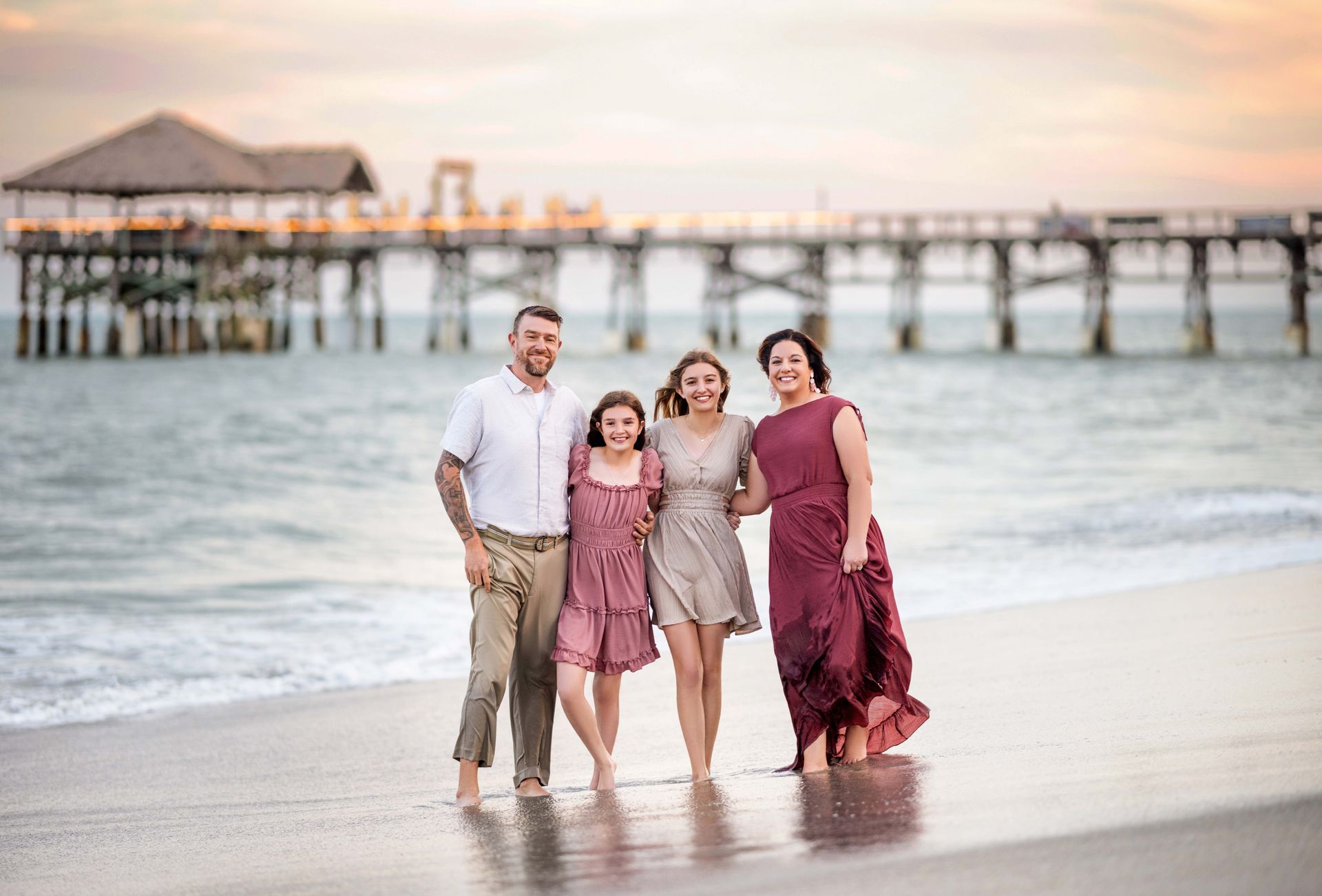 Family of four smiles on a beach with a pier in the background. Sunset colors.