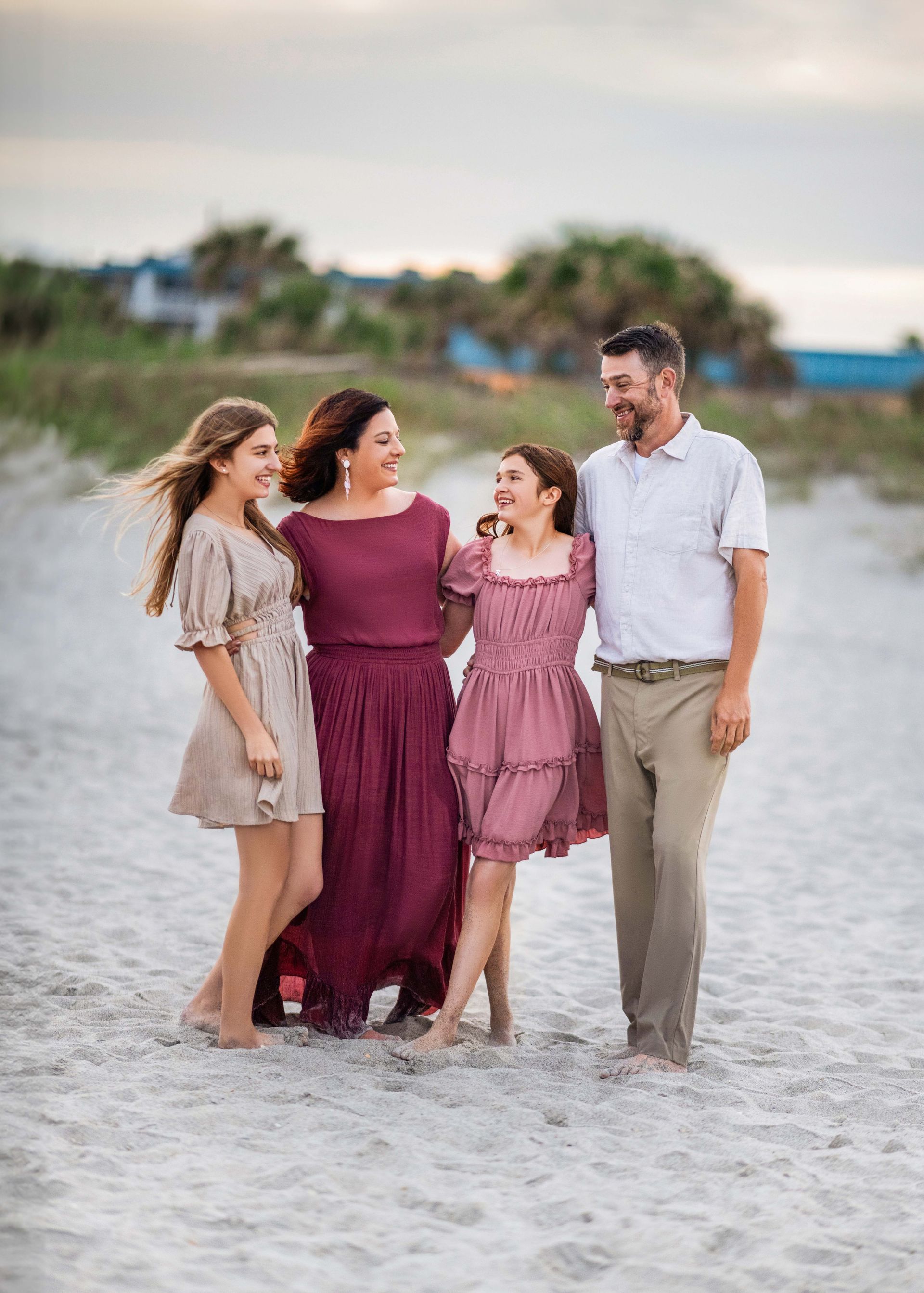 Family of four on a beach, smiling and embracing. Two daughters, a woman in a long dress, and a man in light colors.