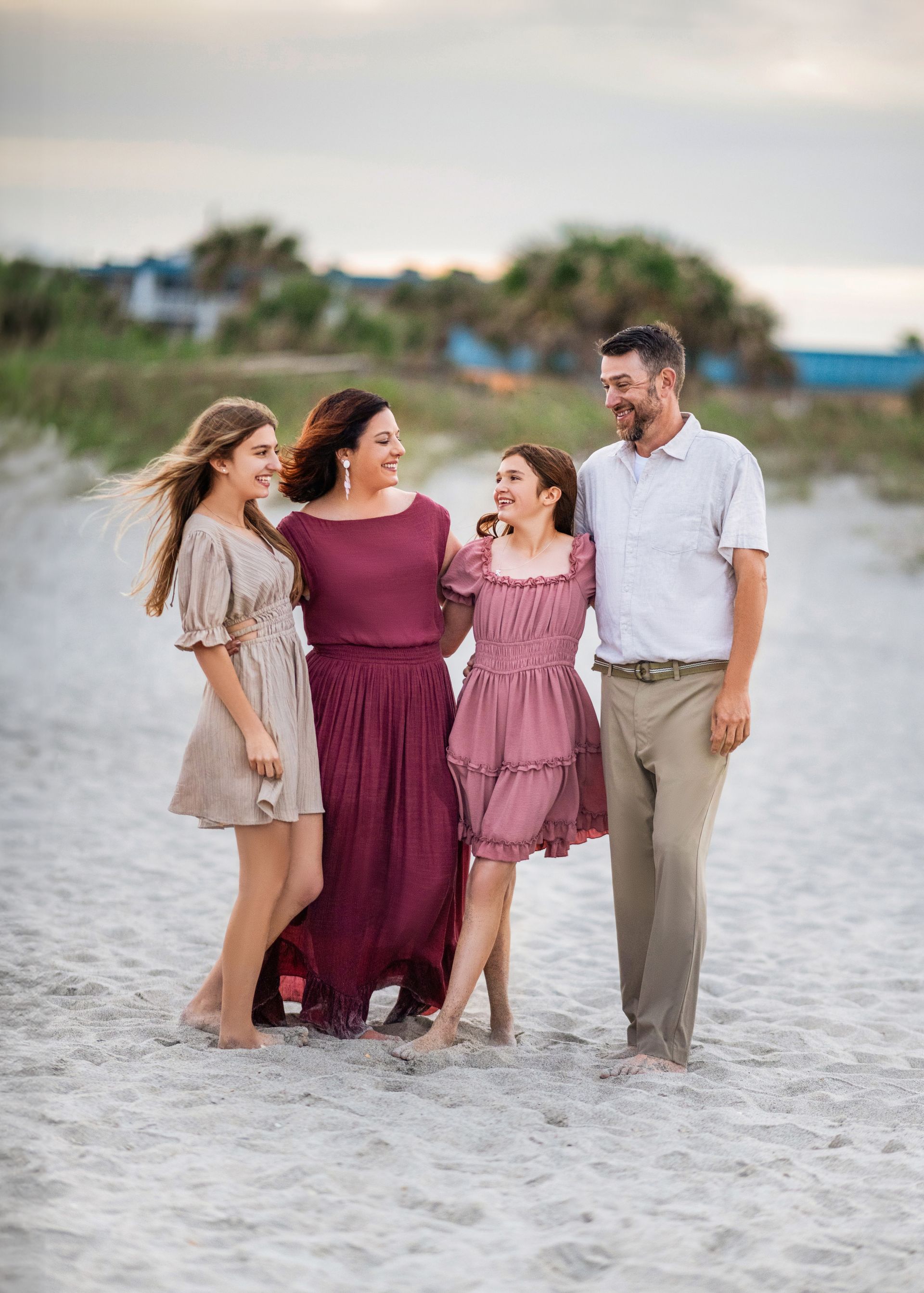 Family of four smiling and hugging on a sandy beach. Sunset hues, woman in burgundy dress.